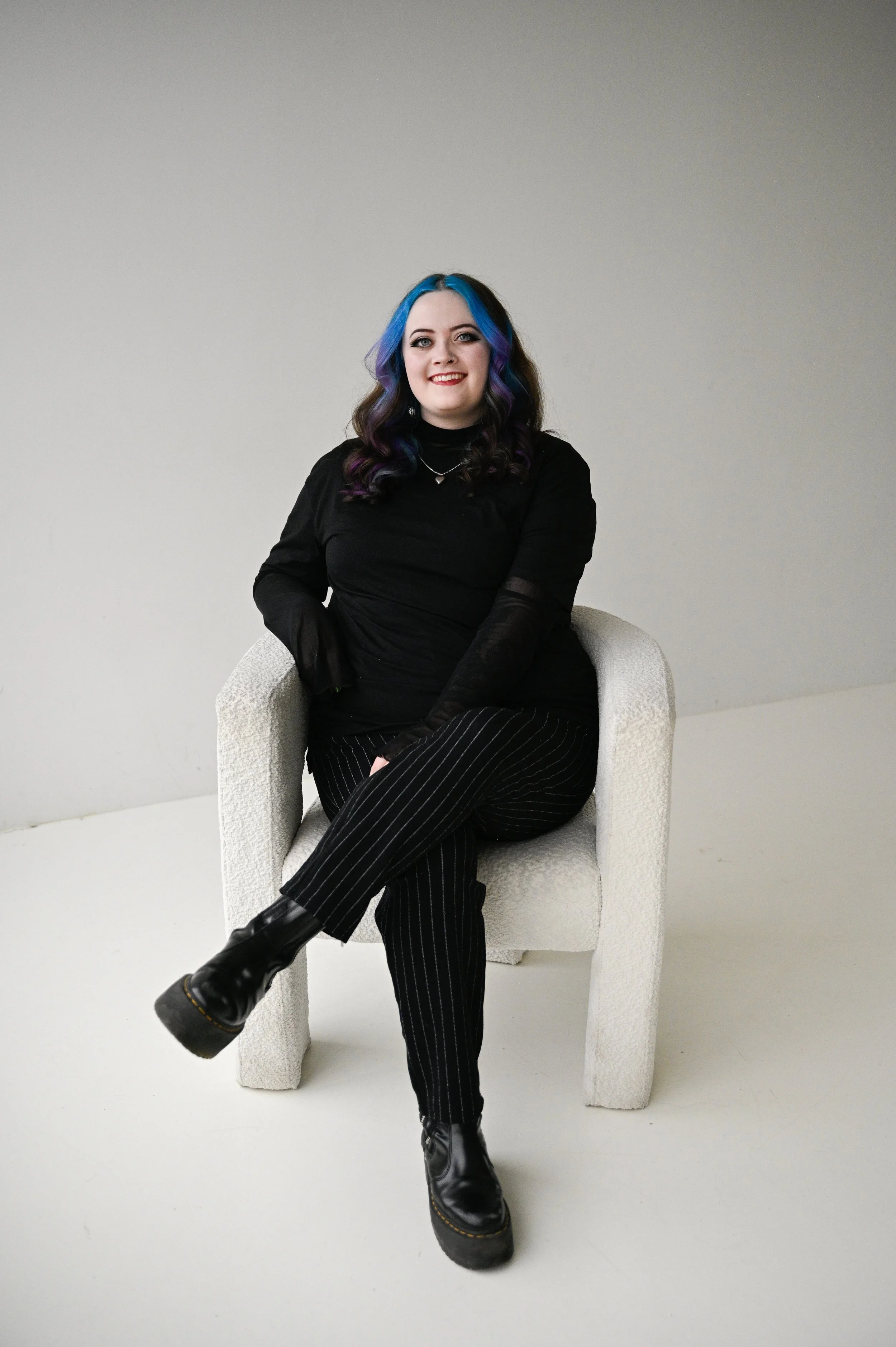 A woman with blue and purple hair sitting on a white textured armchair against a plain light gray background, wearing a black long sleeve top, black striped pants, and black boots, smiling at the camera.