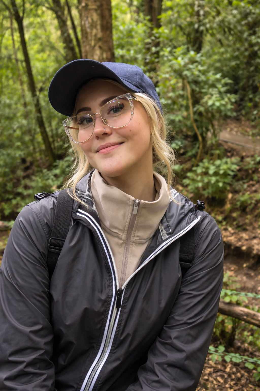 Young woman with glasses and a nose ring smiling in a forest, wearing a navy cap, black jacket, and beige neck warmer.