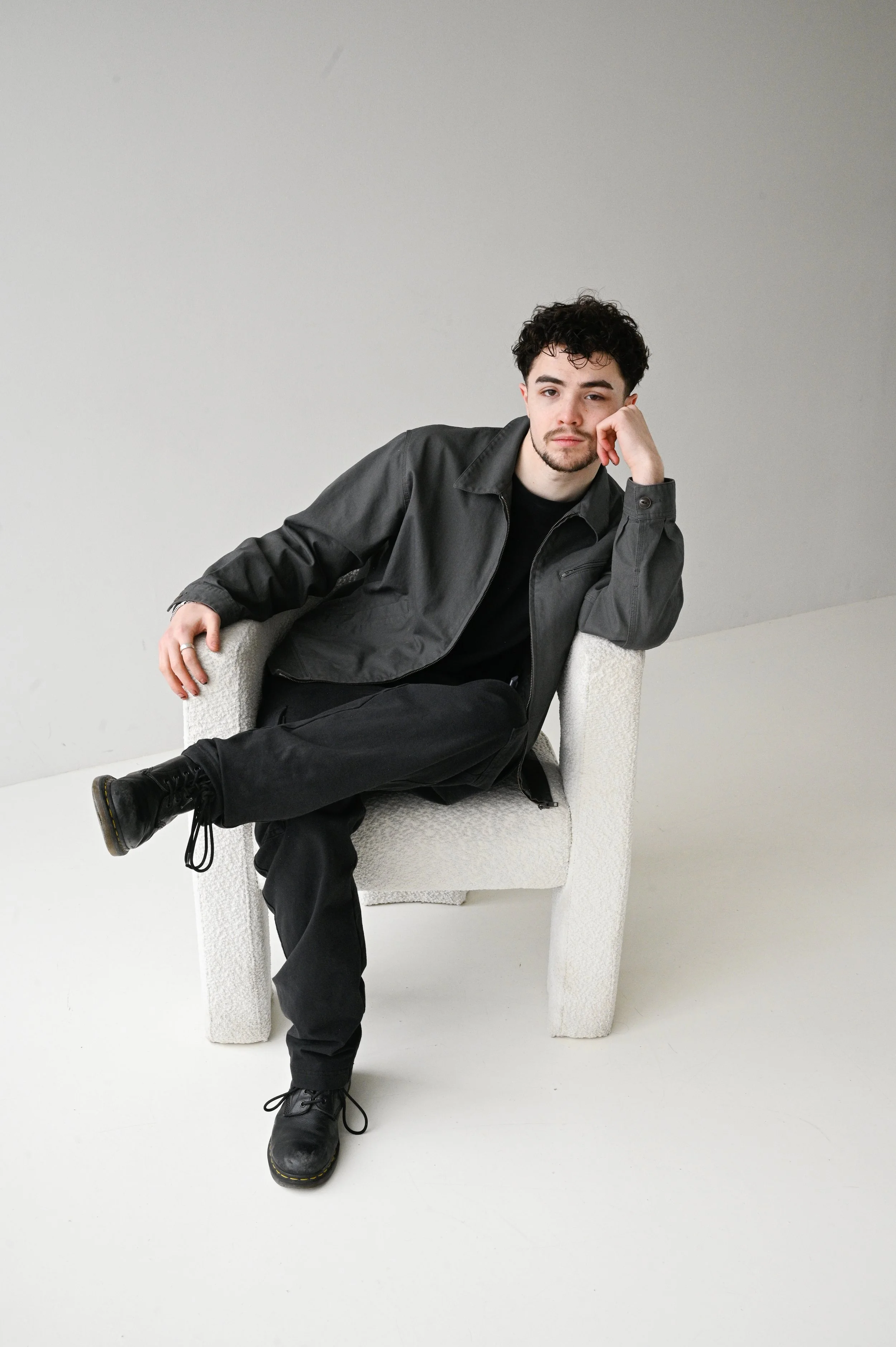 Young man with dark hair, beard, wearing black jacket and shoes, seated casually on a light-colored chair in a minimalistic room with plain gray wall background.
