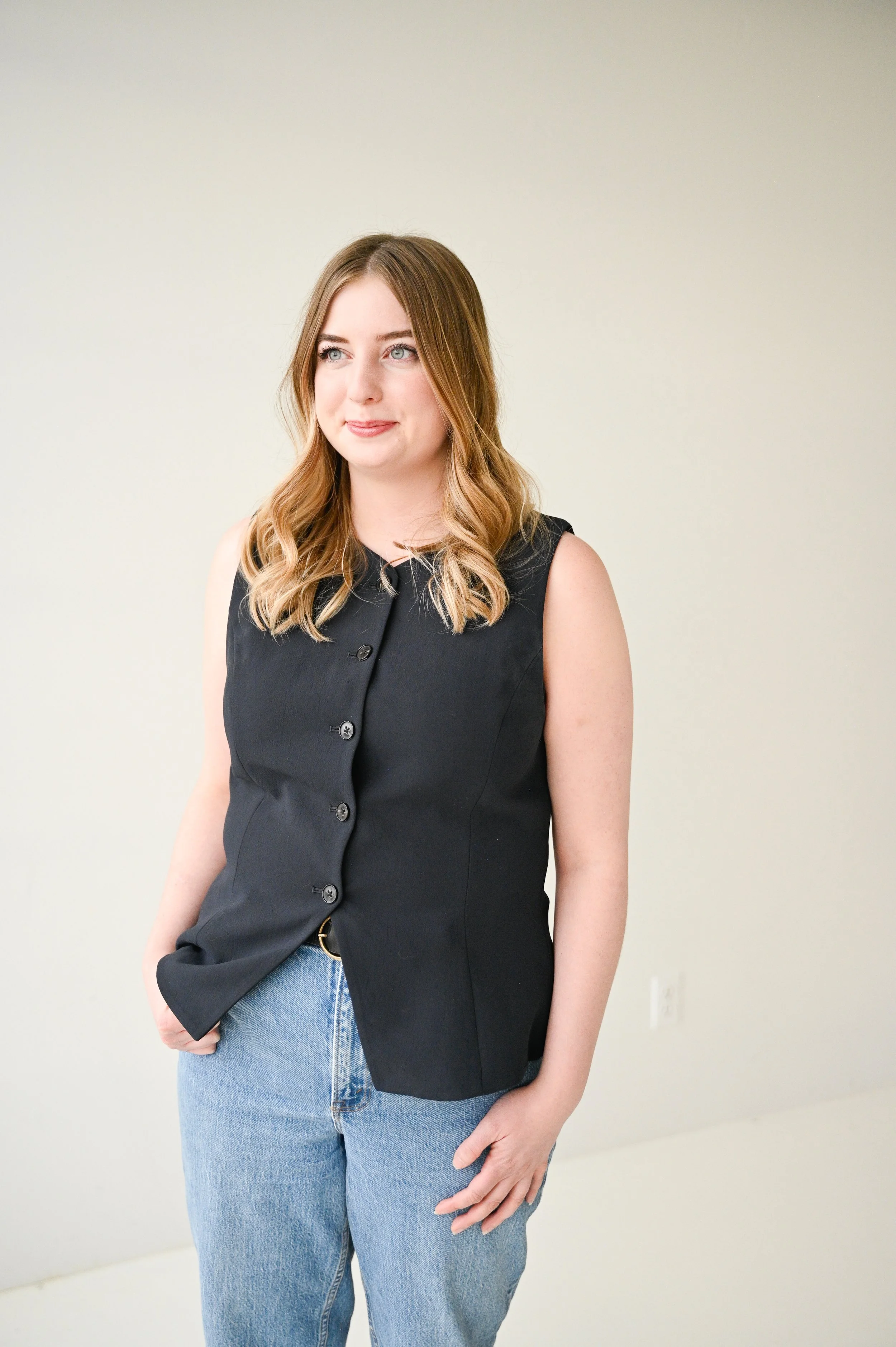 A young woman with shoulder-length blonde hair, wearing a sleeveless black vest and light blue jeans, standing against a plain light-colored background.