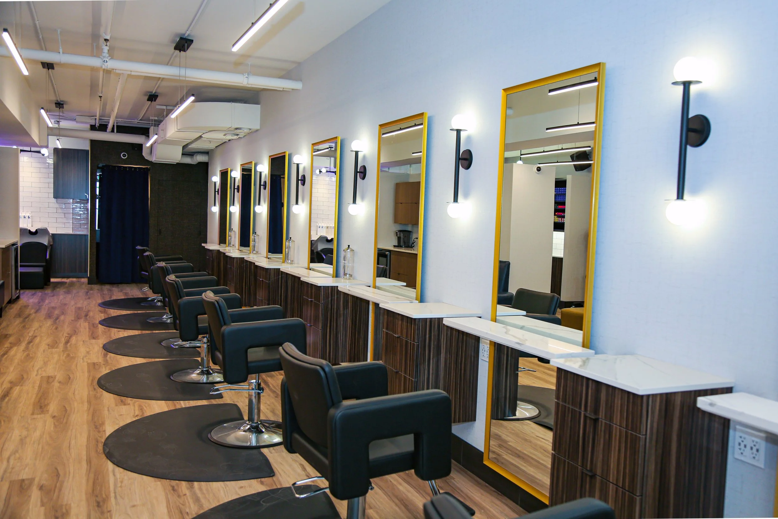 Empty salon with black styling chairs, mirrors with yellow frames, and modern black wall lights, wood flooring, and a white ceiling with exposed pipes.