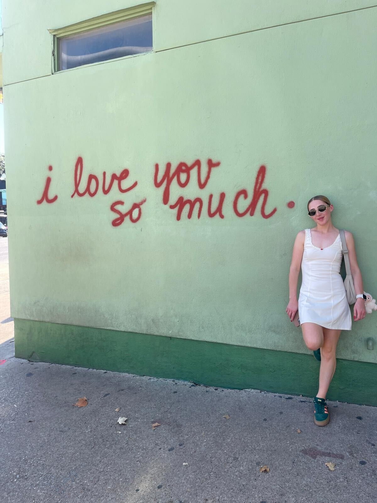 A woman in a white dress wearing sunglasses leaning against a light green wall with red cursive graffiti that reads, 'I love you so much.'