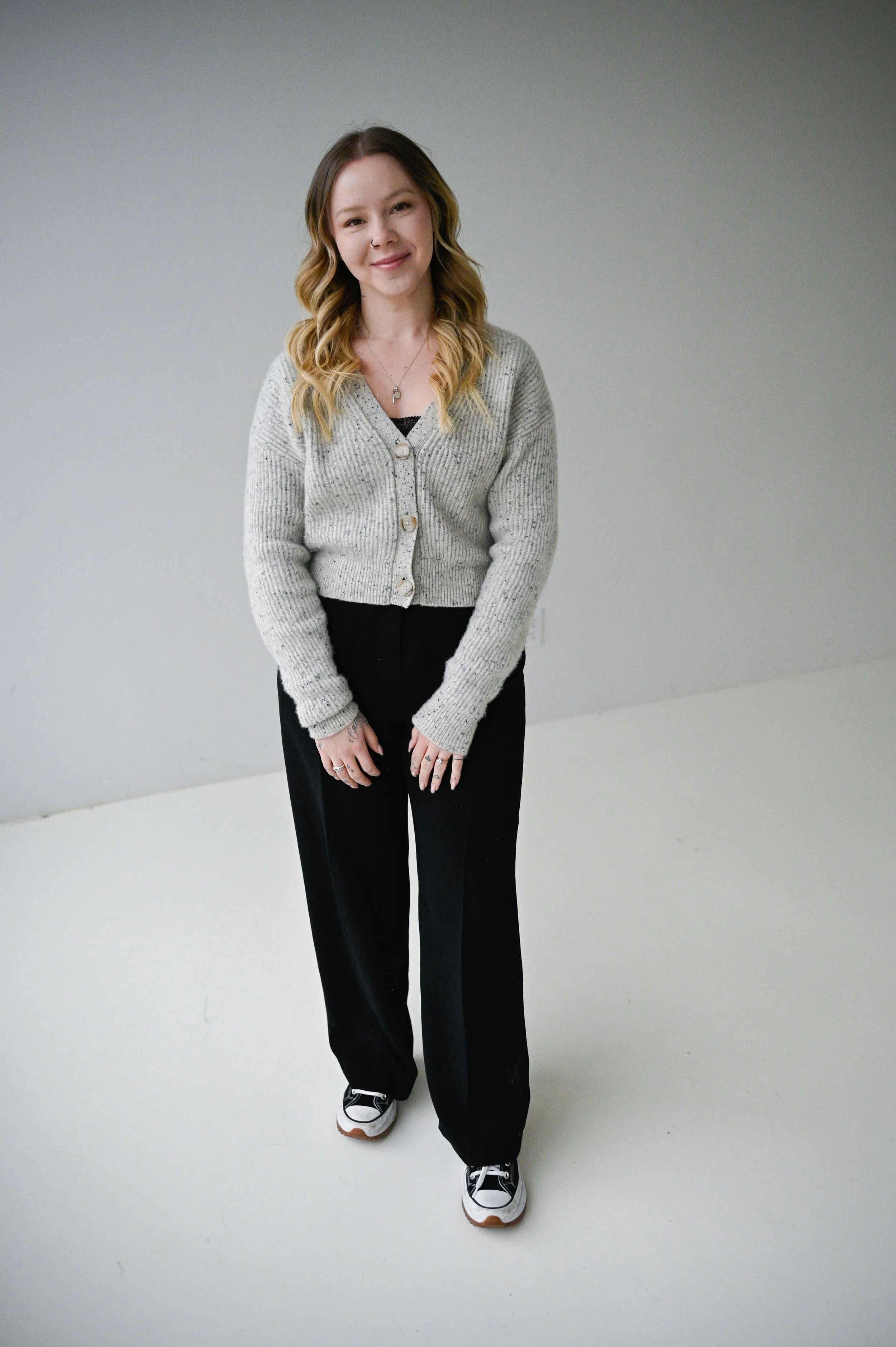 A woman with wavy blonde hair, wearing a gray cardigan, black pants, and sneakers, standing against a plain light gray background and smiling.