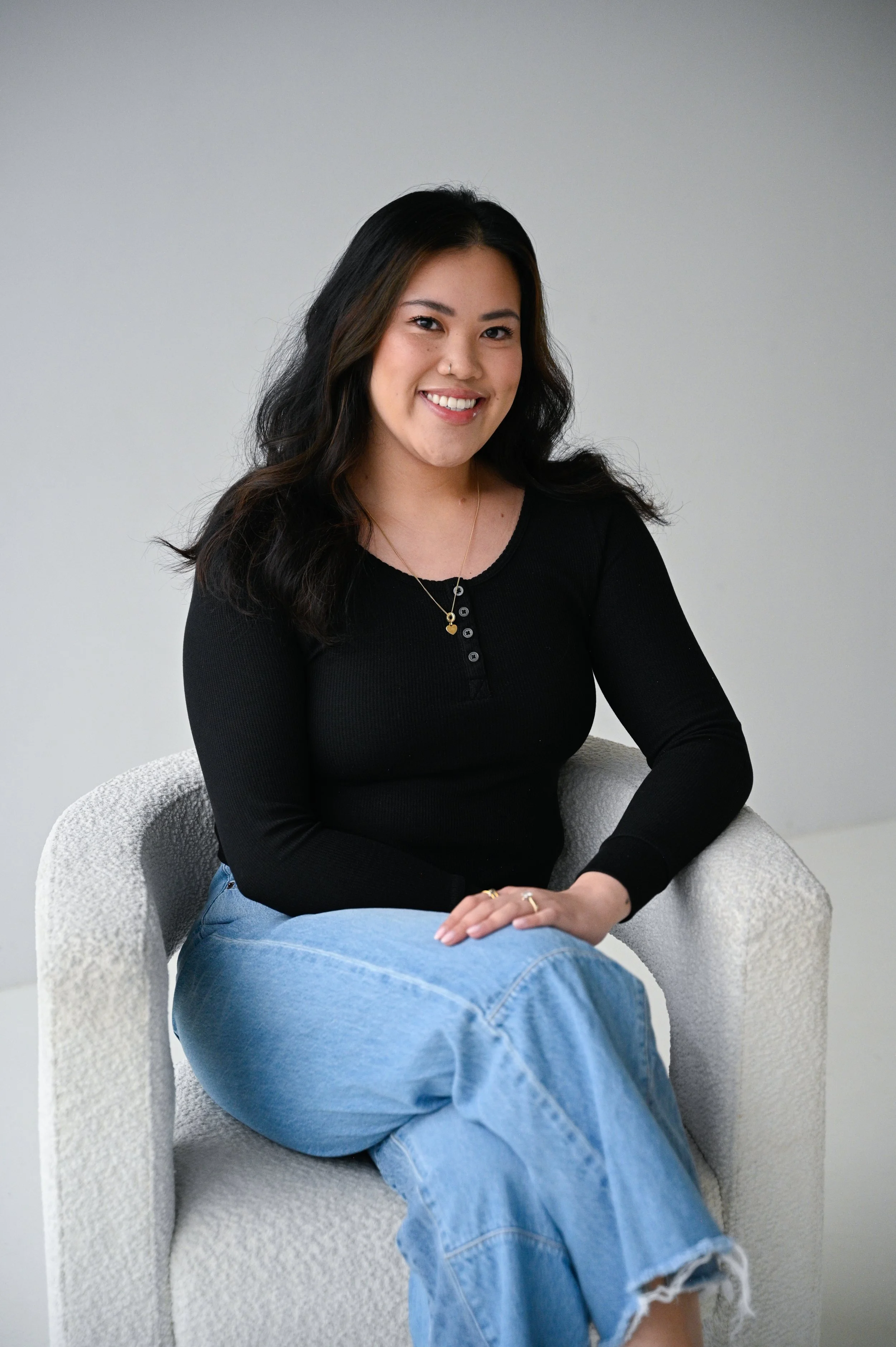 A young woman with black hair, wearing a black long-sleeve top and light blue jeans, sitting on a white upholstered chair against a plain light gray wall, smiling at the camera.