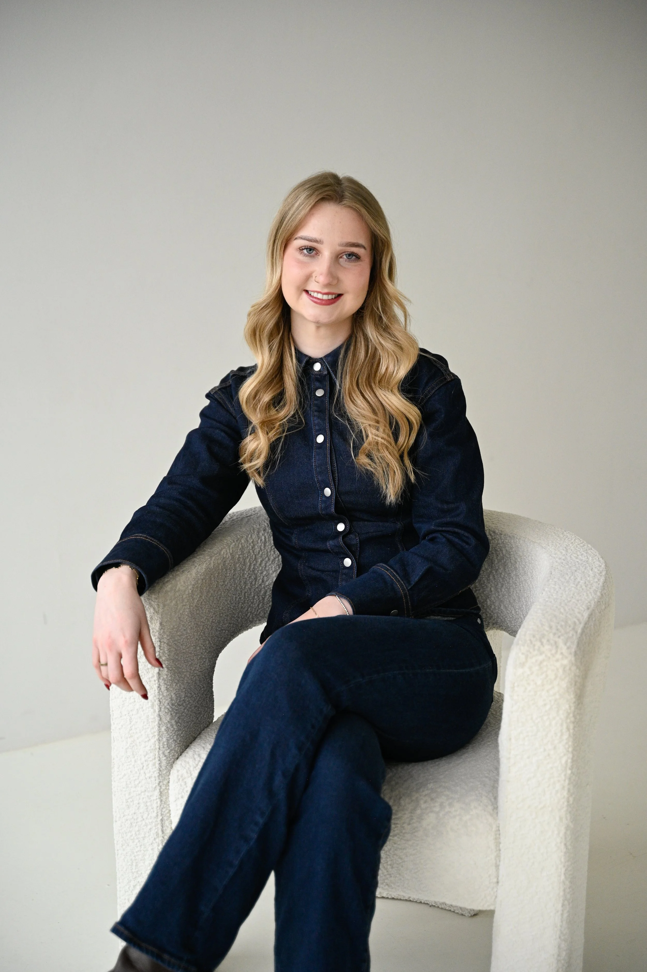 Young woman with long blonde hair smiling, sitting in a beige armchair, wearing a dark denim shirt and jeans, against a plain light gray wall.