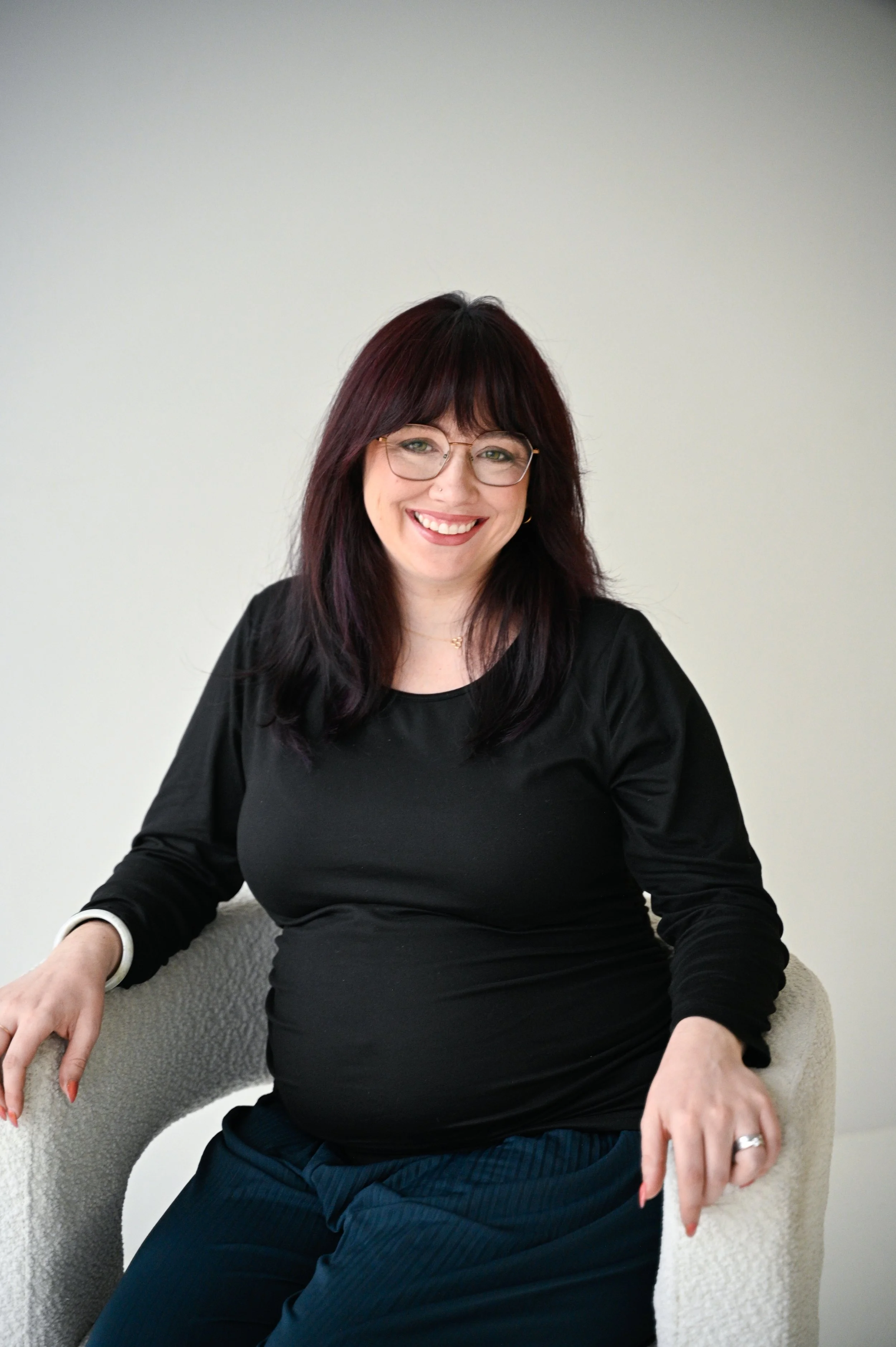 A woman with dark red hair, glasses, and a wide smile sitting on a beige textured armchair, wearing a black long-sleeve top and navy blue pants, in front of a plain off-white wall.