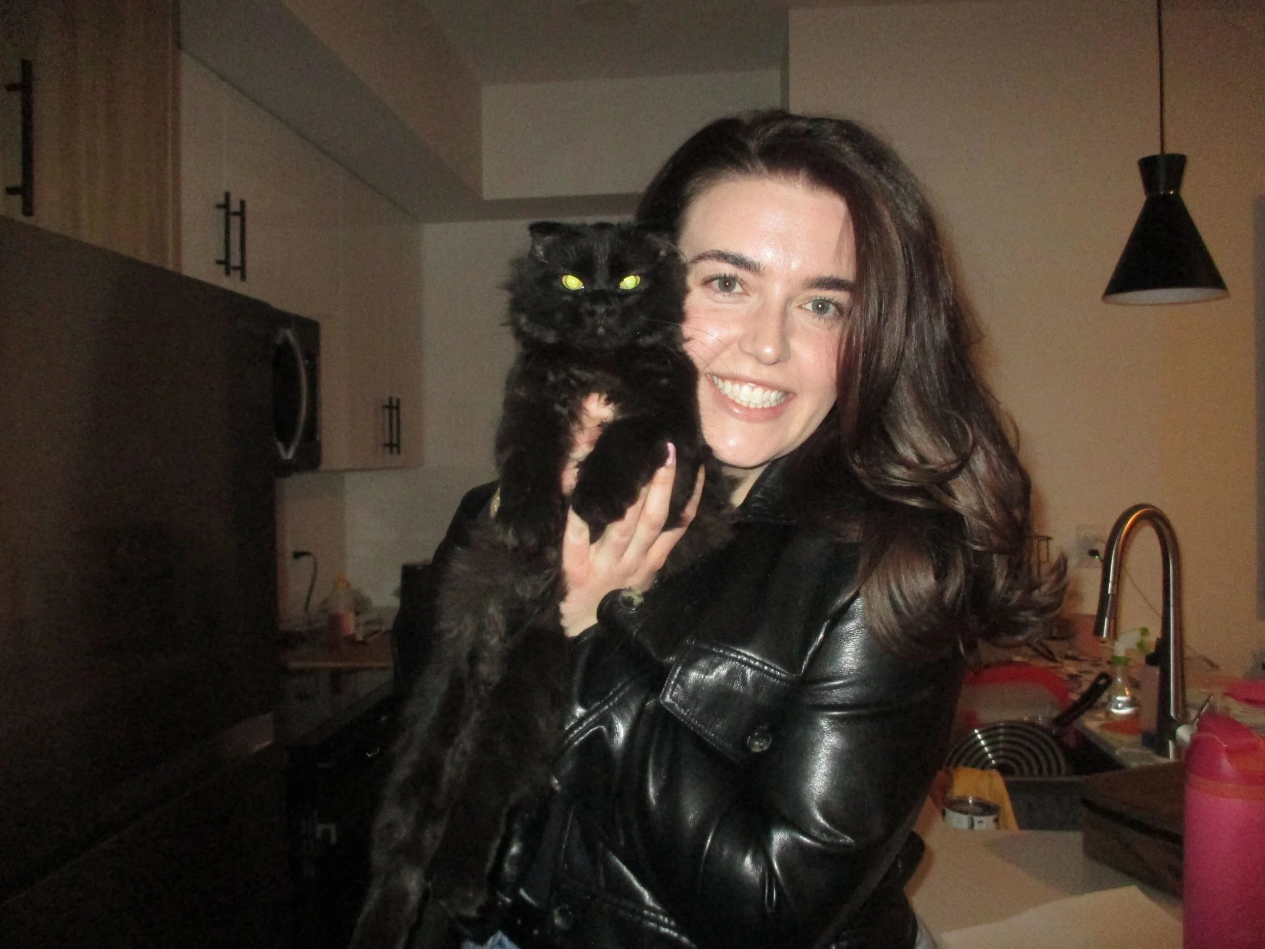 A woman with long brown hair and a black leather jacket holding a black cat in a kitchen.