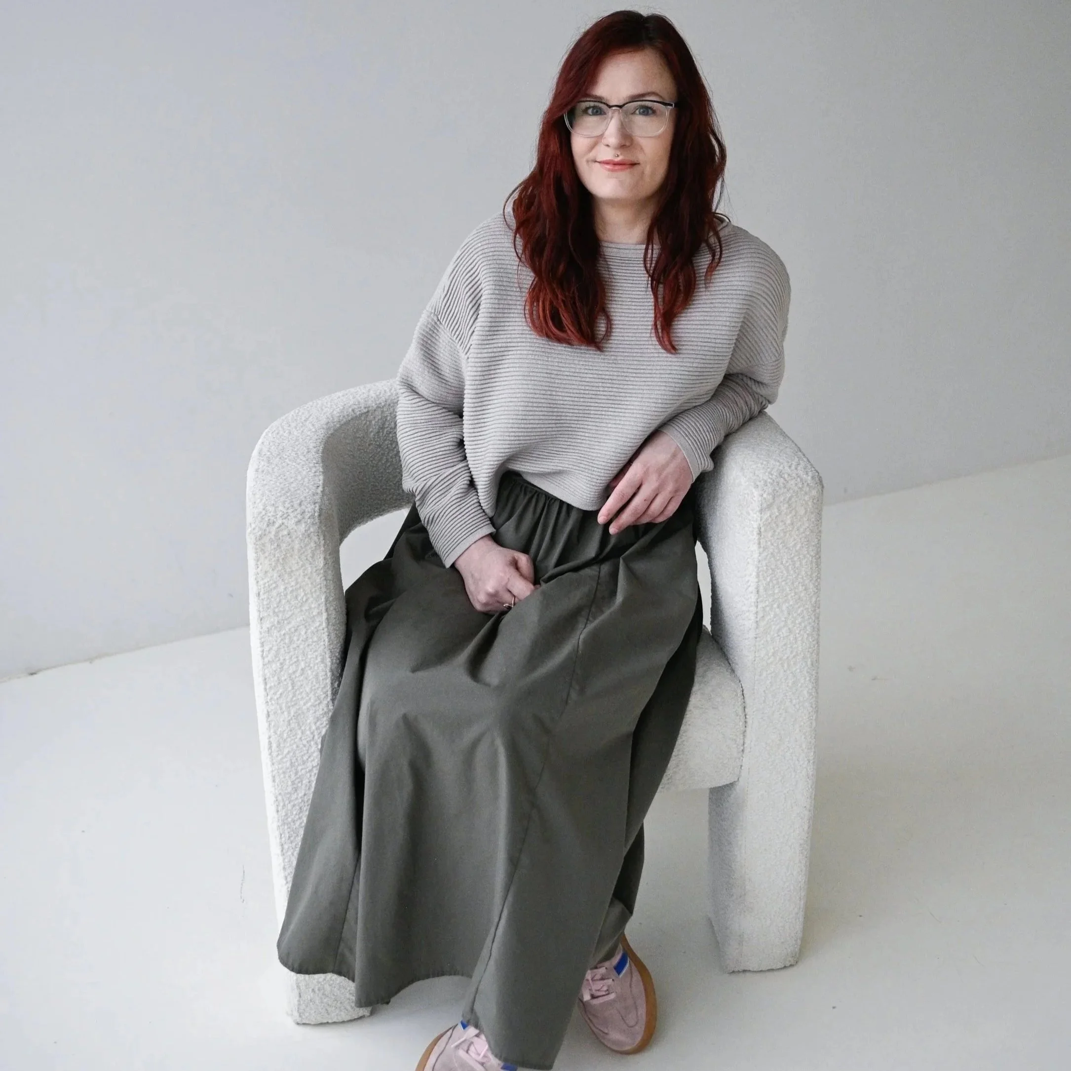 Woman with red hair and glasses sitting on white armchair, wearing a beige sweater, dark skirt, and pink sneakers.