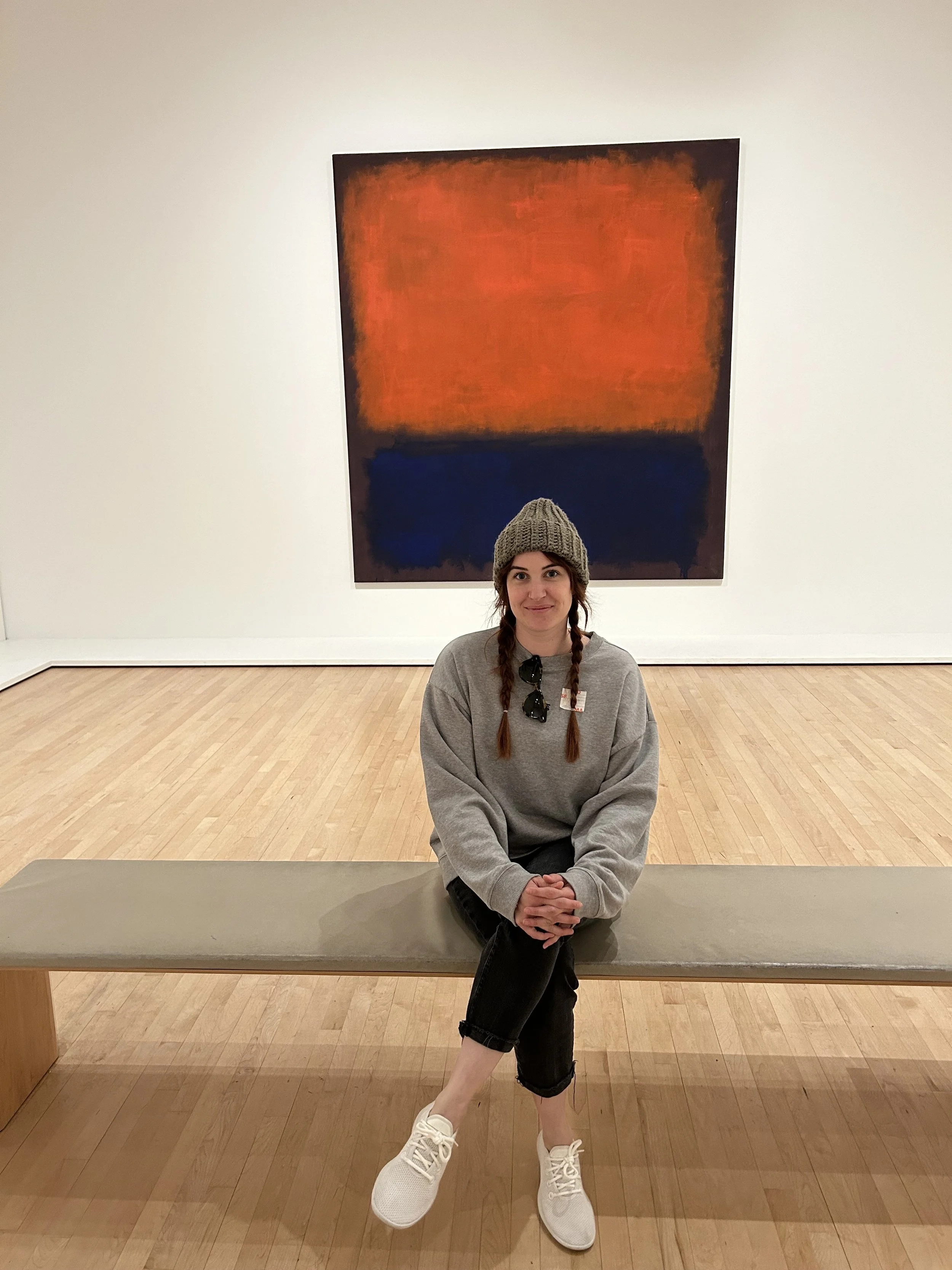A young woman with braided hair, wearing a gray sweater, black pants, white sneakers, and a knit hat, sitting on a bench in an art gallery with abstract artwork in the background.