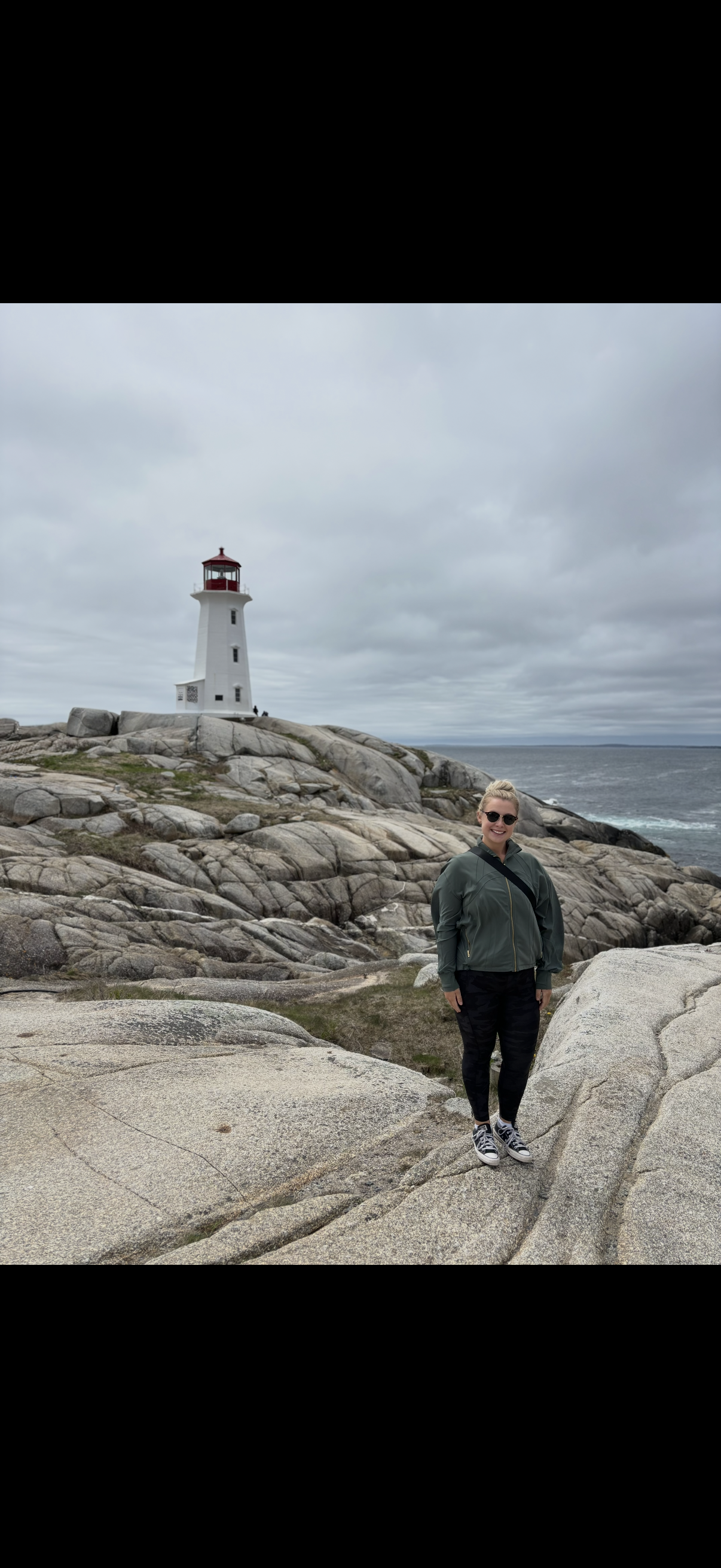 A woman with blonde hair, wearing sunglasses, a green jacket, and black pants, standing on rocky terrain near the coast with a lighthouse in the background under a cloudy sky.