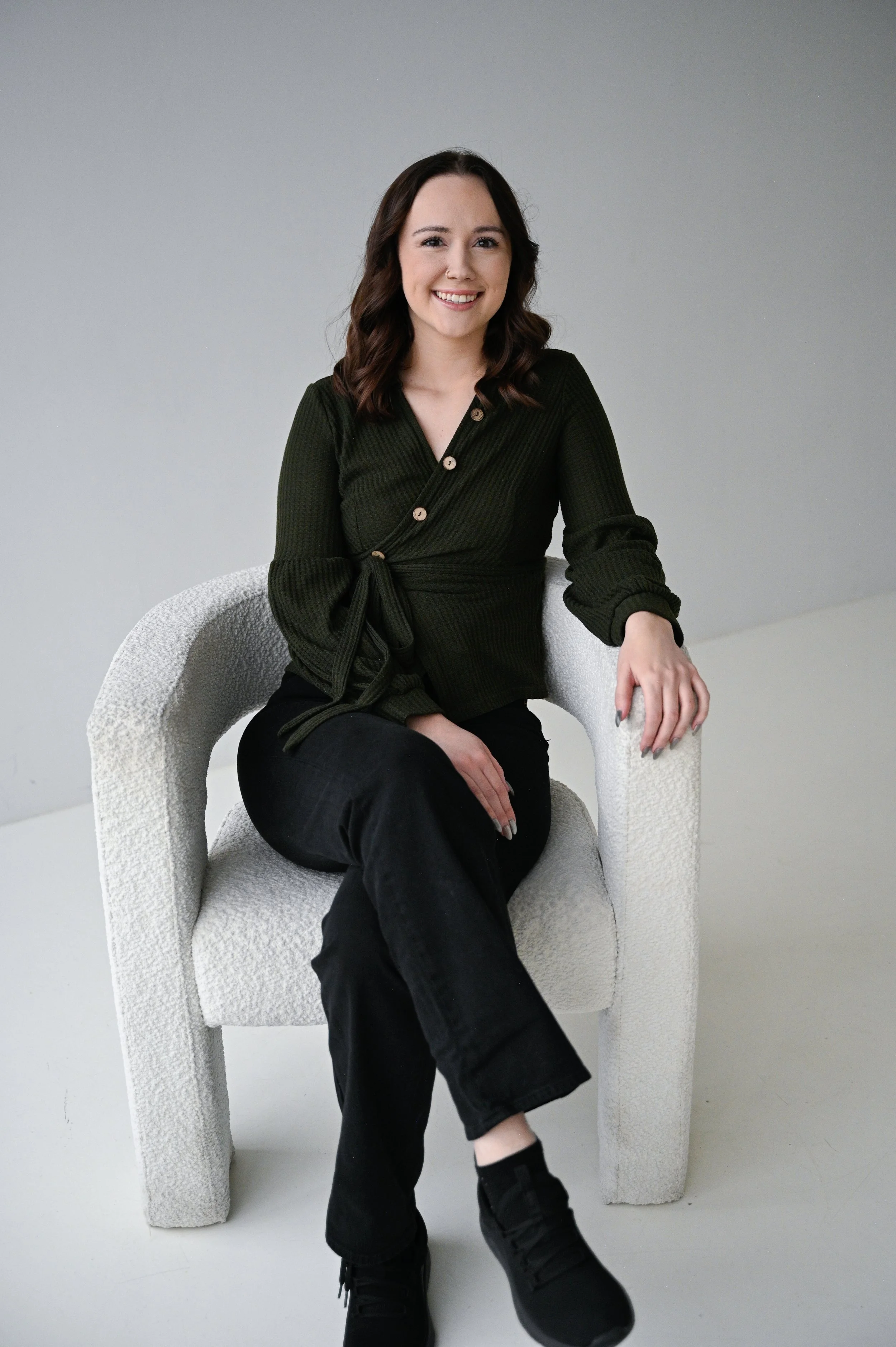 A woman with shoulder-length brown hair smiling, sitting on a textured armchair against a plain gray background, wearing a dark green wrap-style blouse and black pants.
