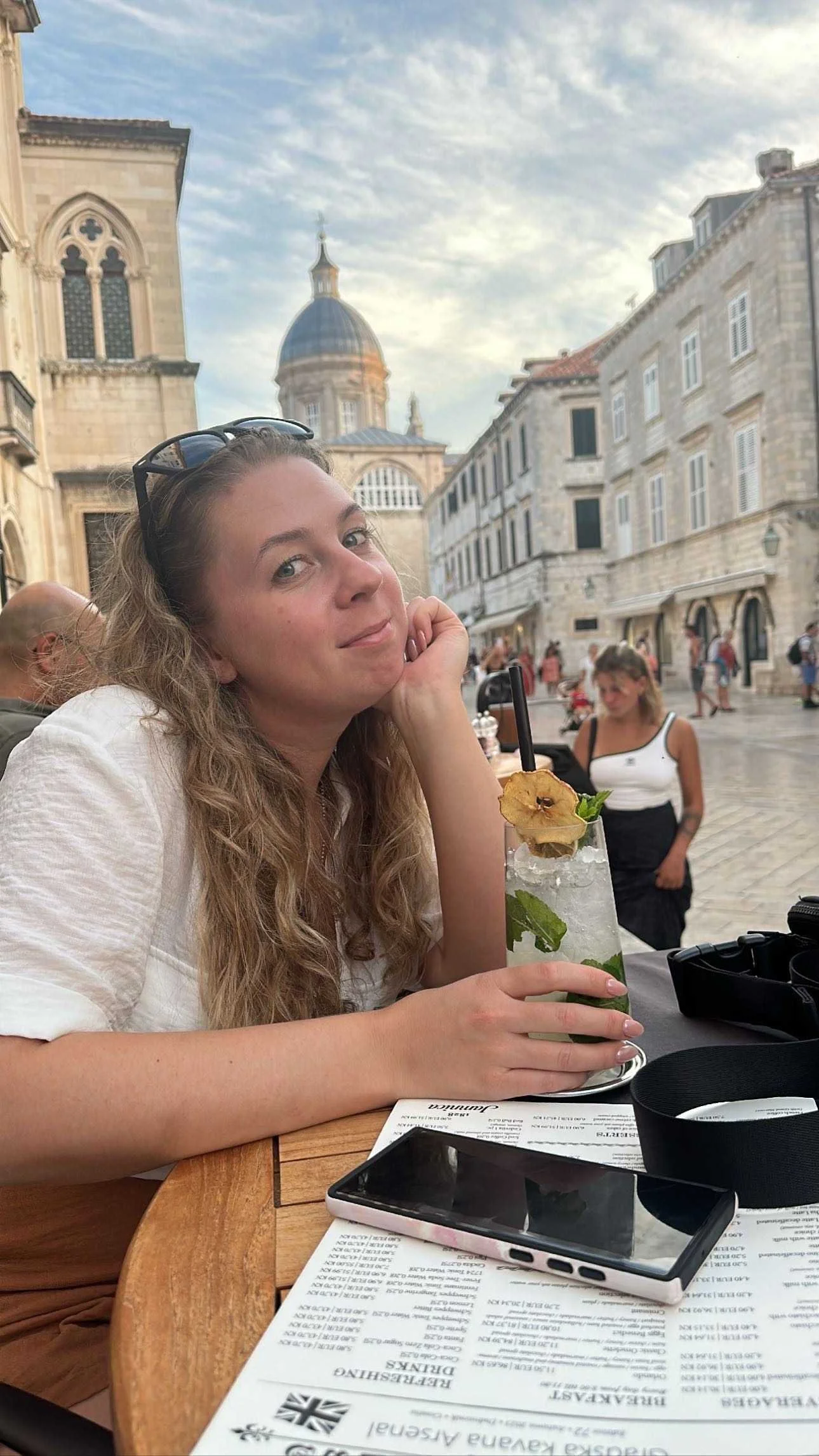 A woman with long curly hair sitting at an outdoor restaurant table, resting her chin on her hand, with a tall drink garnished with a dried apple slice and mint leaves. In the background are historic European-style buildings and a dome.
