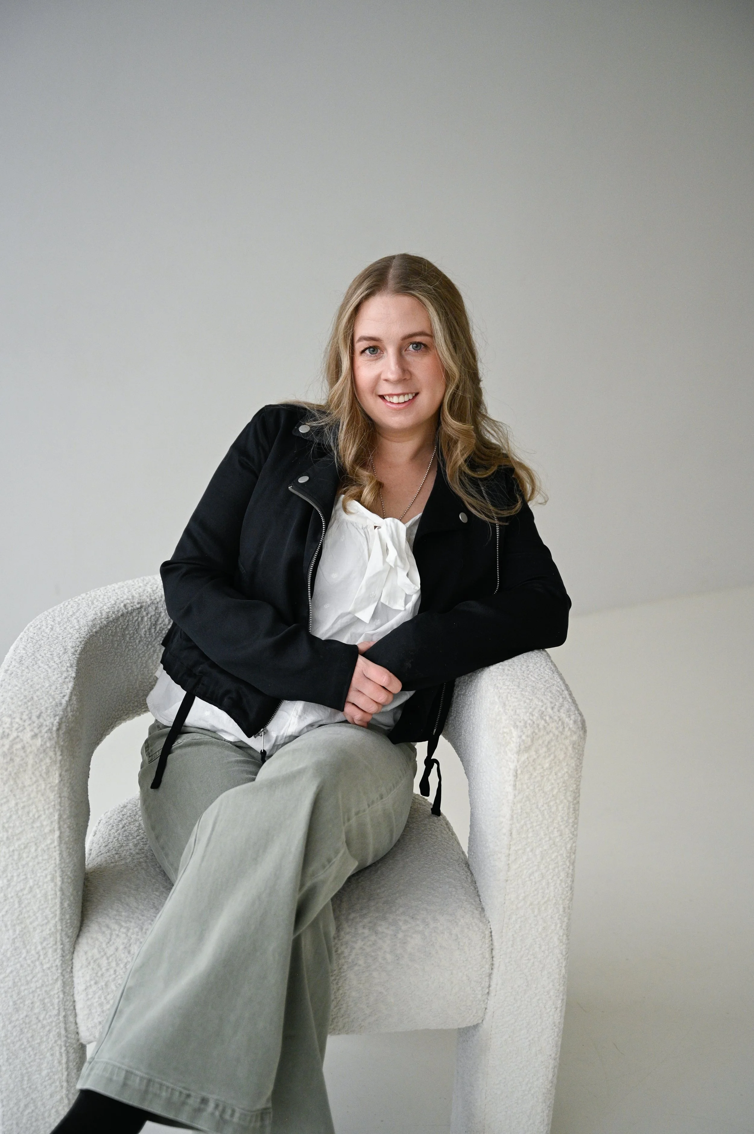 Young woman sitting on a textured white armchair, smiling at the camera, wearing a black jacket, white blouse, and light-colored pants, against a plain light background.