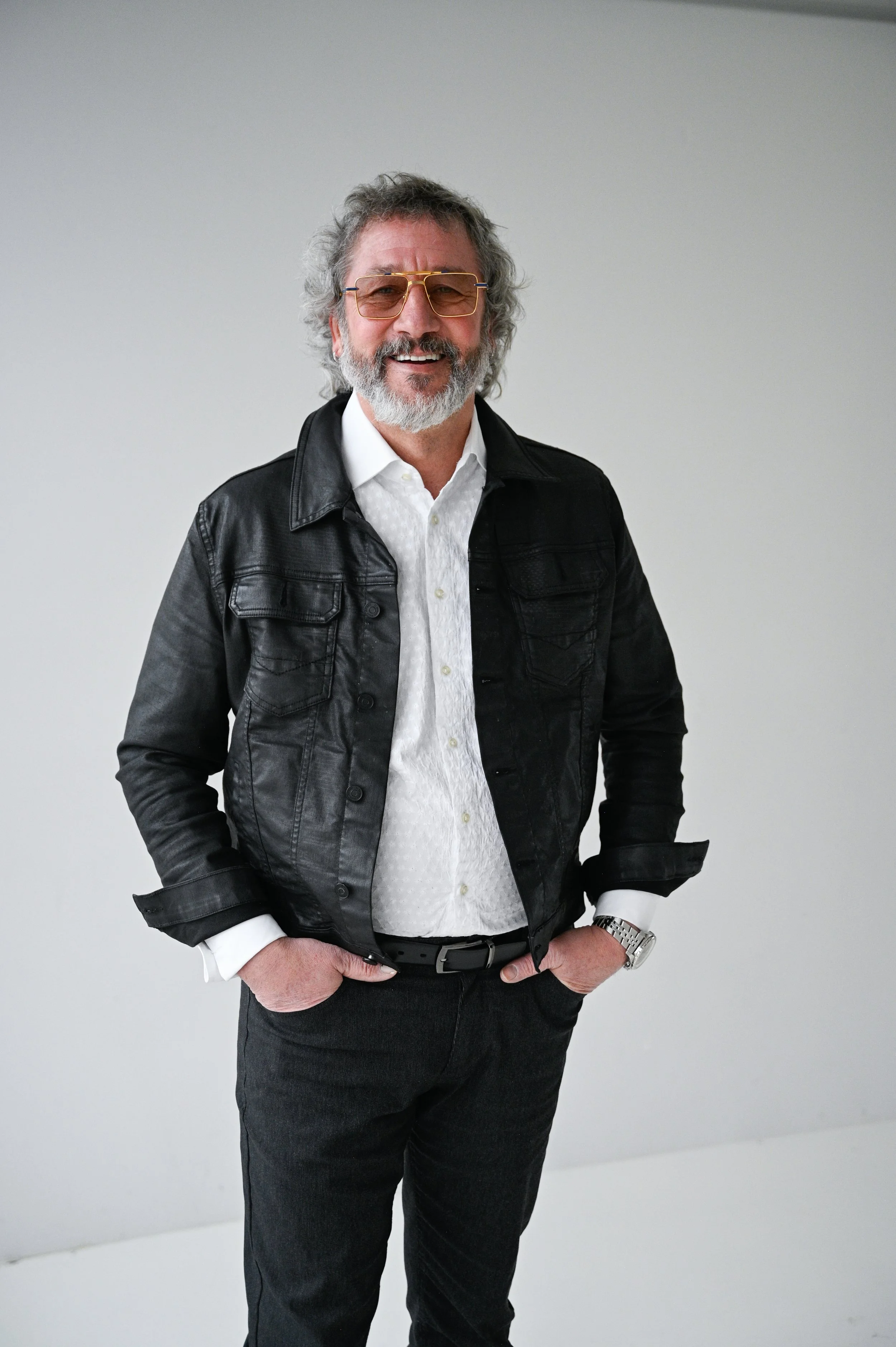 A man with gray hair and a beard, wearing sunglasses, a black leather jacket, white shirt, and dark pants, smiling and standing with hands in pockets against a plain light gray background.