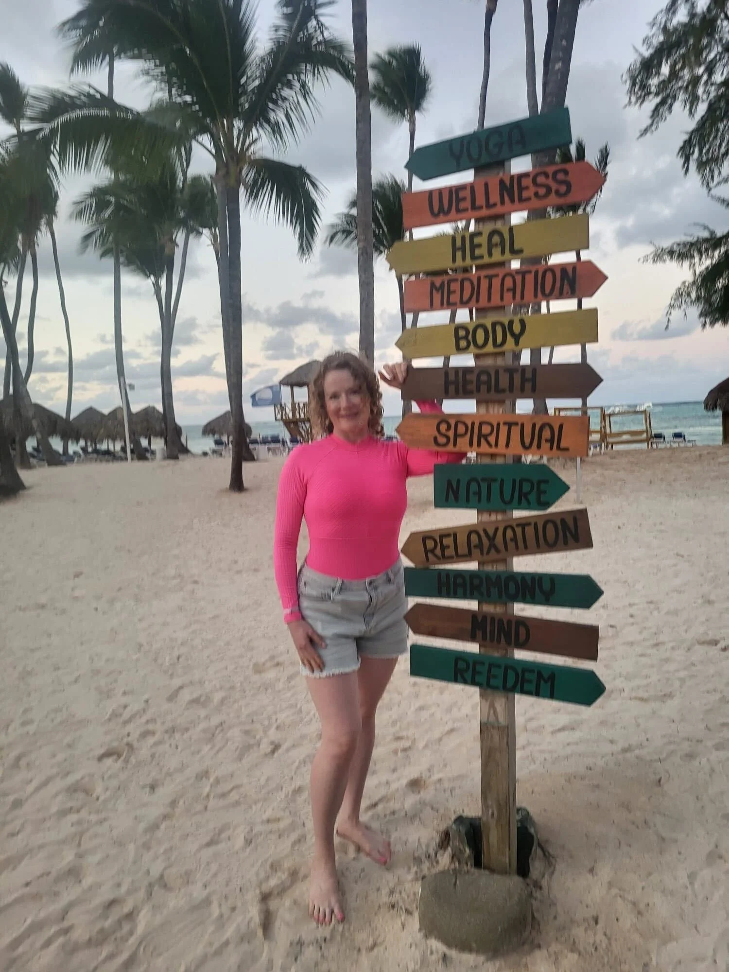 Woman in pink long sleeve top and denim shorts standing next to a colorful directional signs on the beach with palm trees and huts in the background.