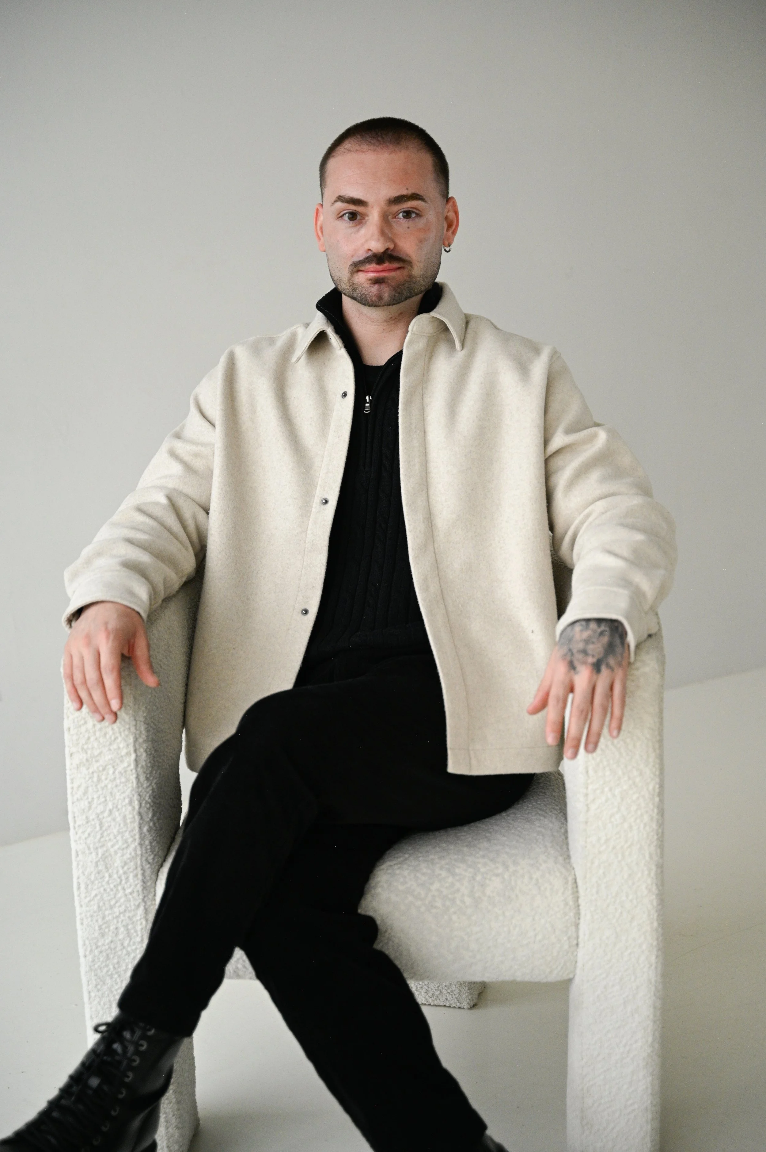 Man with short hair, beard, and earrings sitting in a cream-colored armchair, wearing a beige jacket, black shirt, black pants, and black boots, with a tattooed hand resting on the armrest.