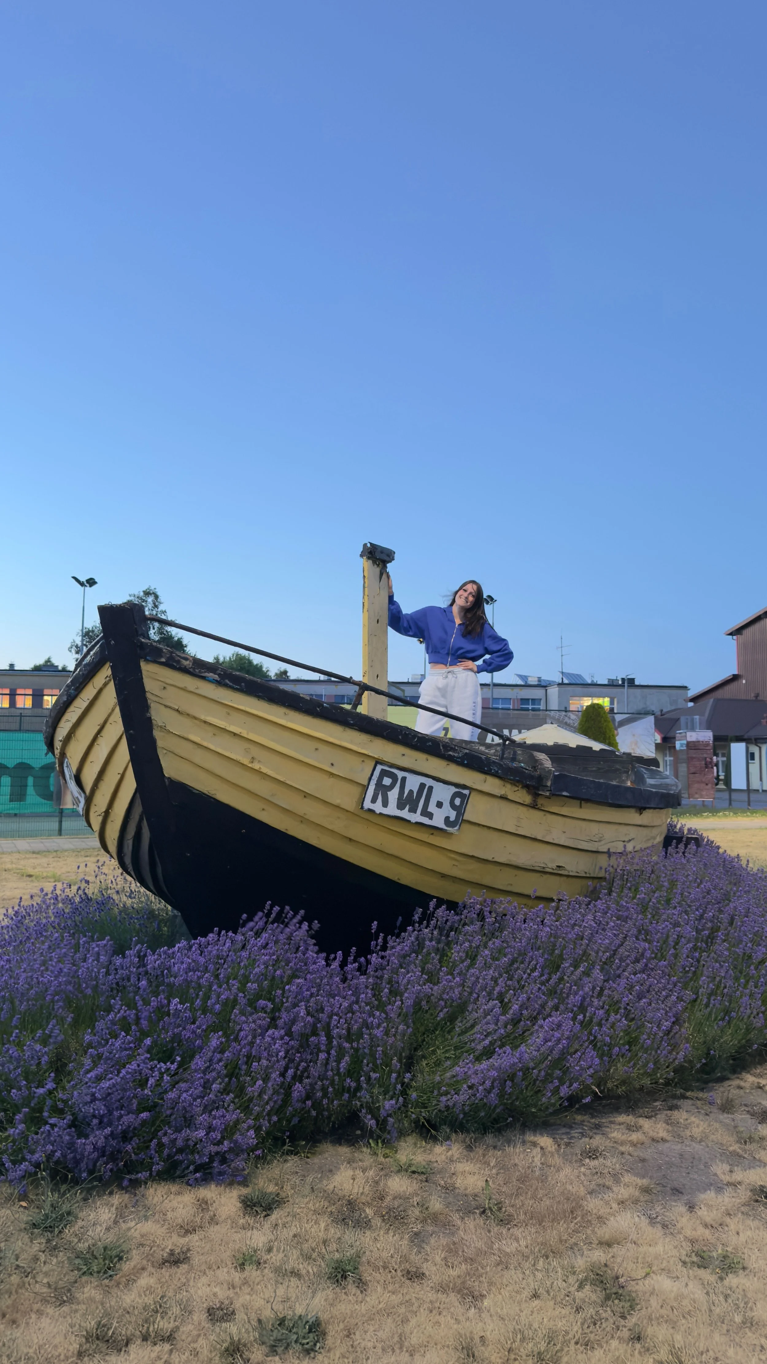 A woman in a blue jacket and white pants standing on a boat-shaped structure surrounded by purple flowers, with a clear blue sky in the background.