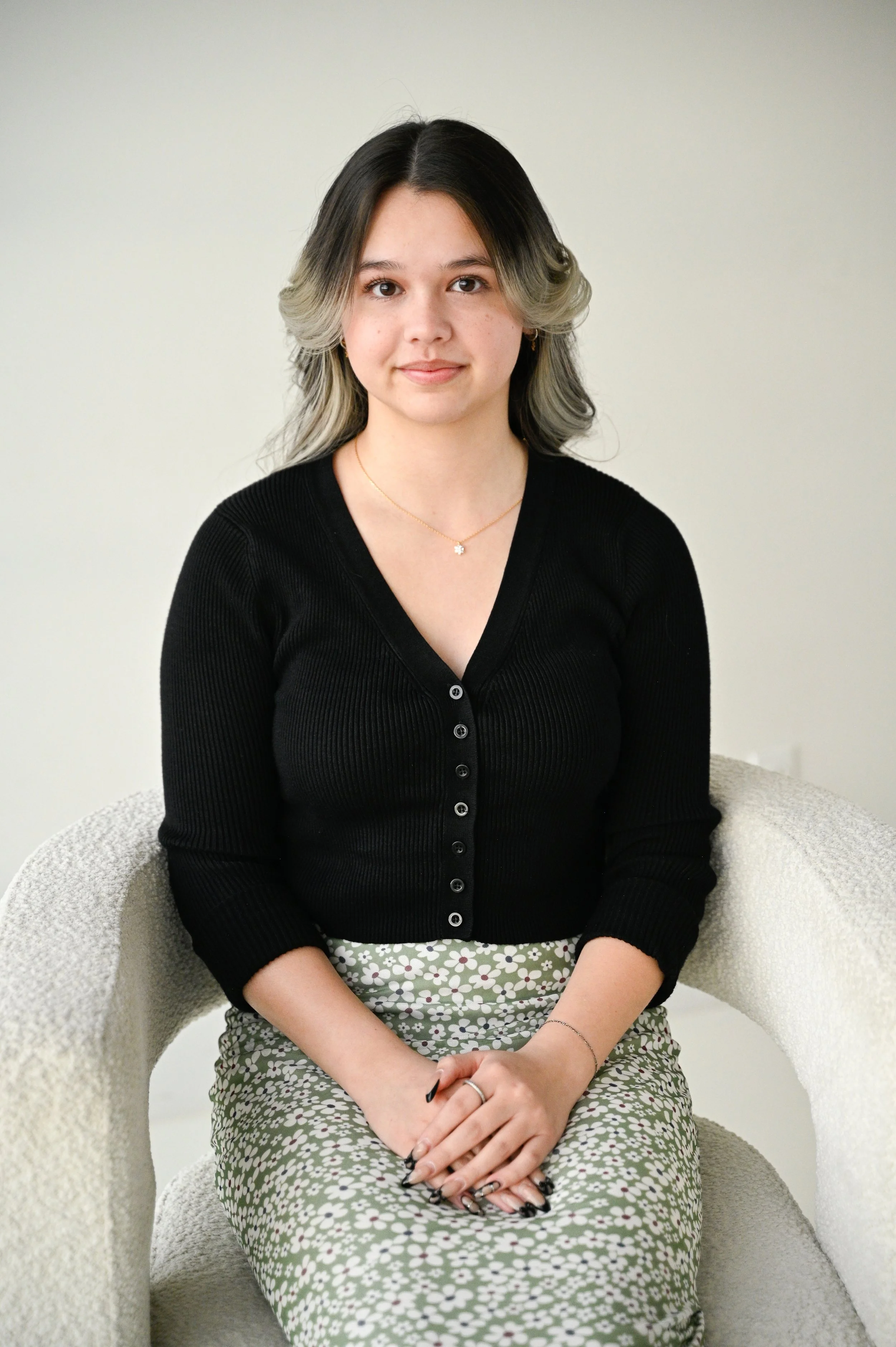 A young woman with shoulder-length, wavy hair with blonde tips, sitting on a light-colored textured chair against a plain off-white background. She is wearing a black button-up top and a light green floral skirt, with her hands resting on her lap.