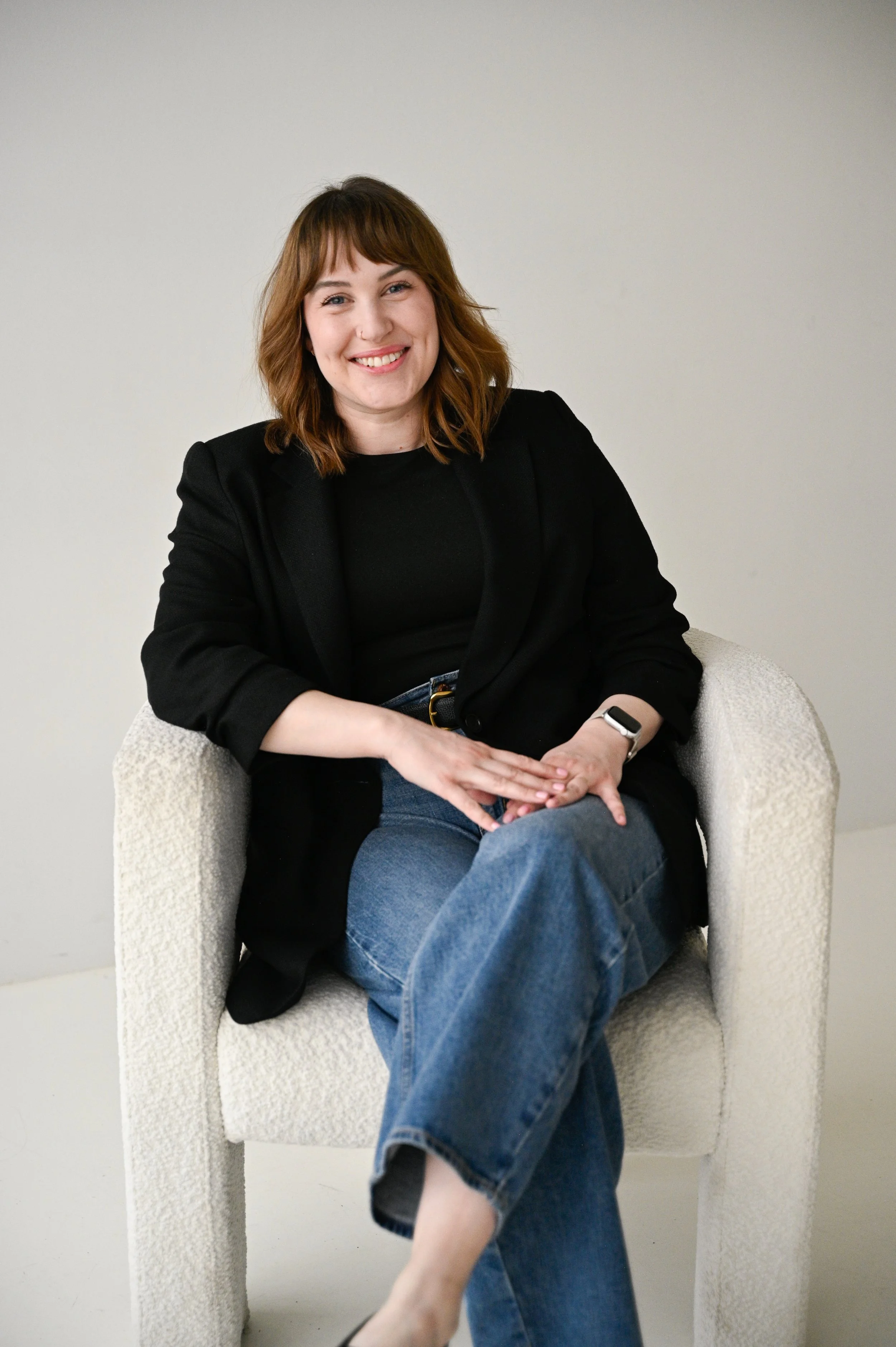 A woman with shoulder-length light brown hair, smiling, sitting in a beige textured armchair against a plain light background. She is wearing a black blazer, black top, blue jeans, and a smartwatch on her left wrist.
