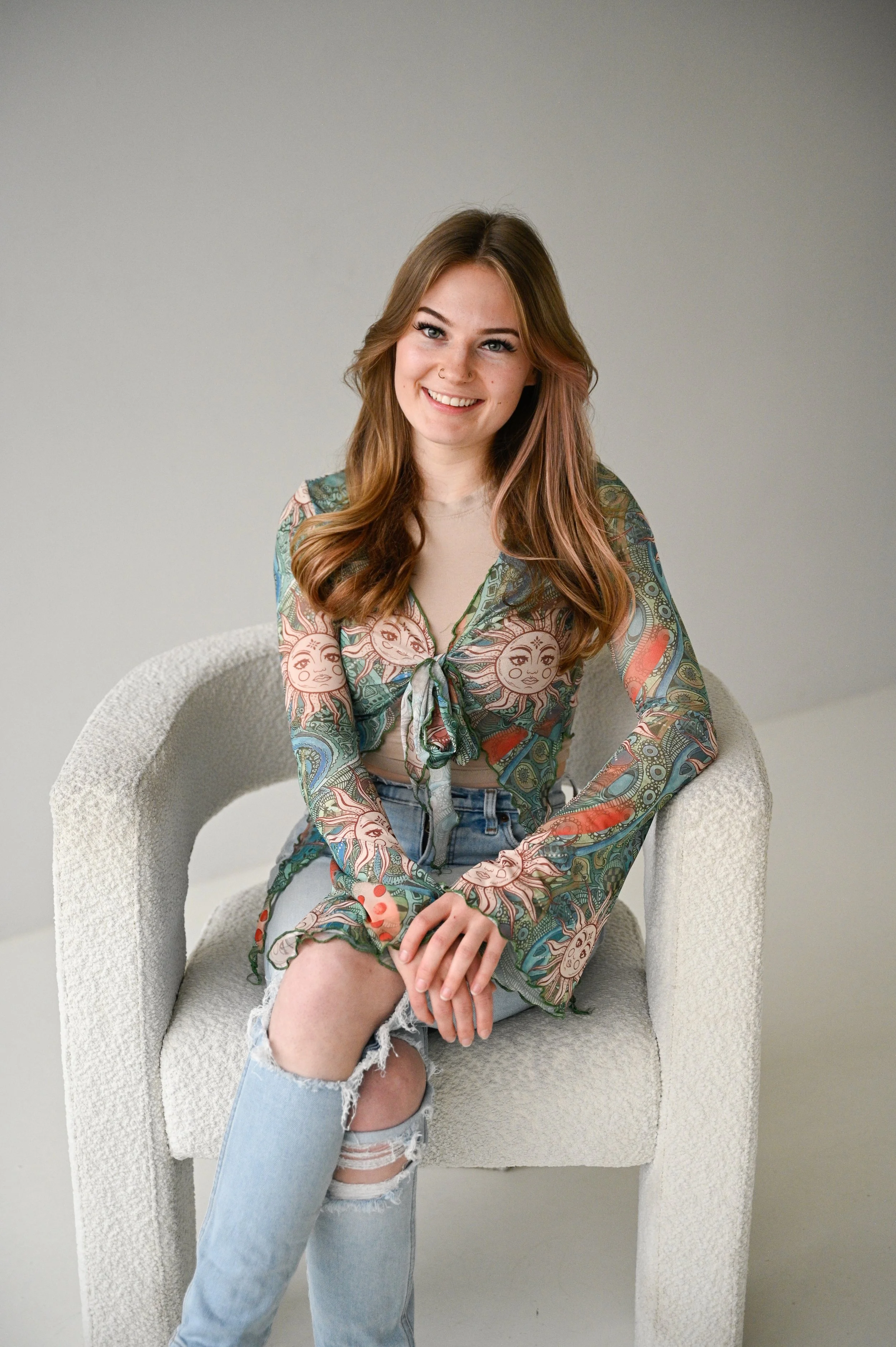 Young woman with long wavy hair sitting on a cream-colored textured armchair, smiling at the camera, wearing a colorful sun and moon patterned sheer blouse, a beige top underneath, and ripped jeans.