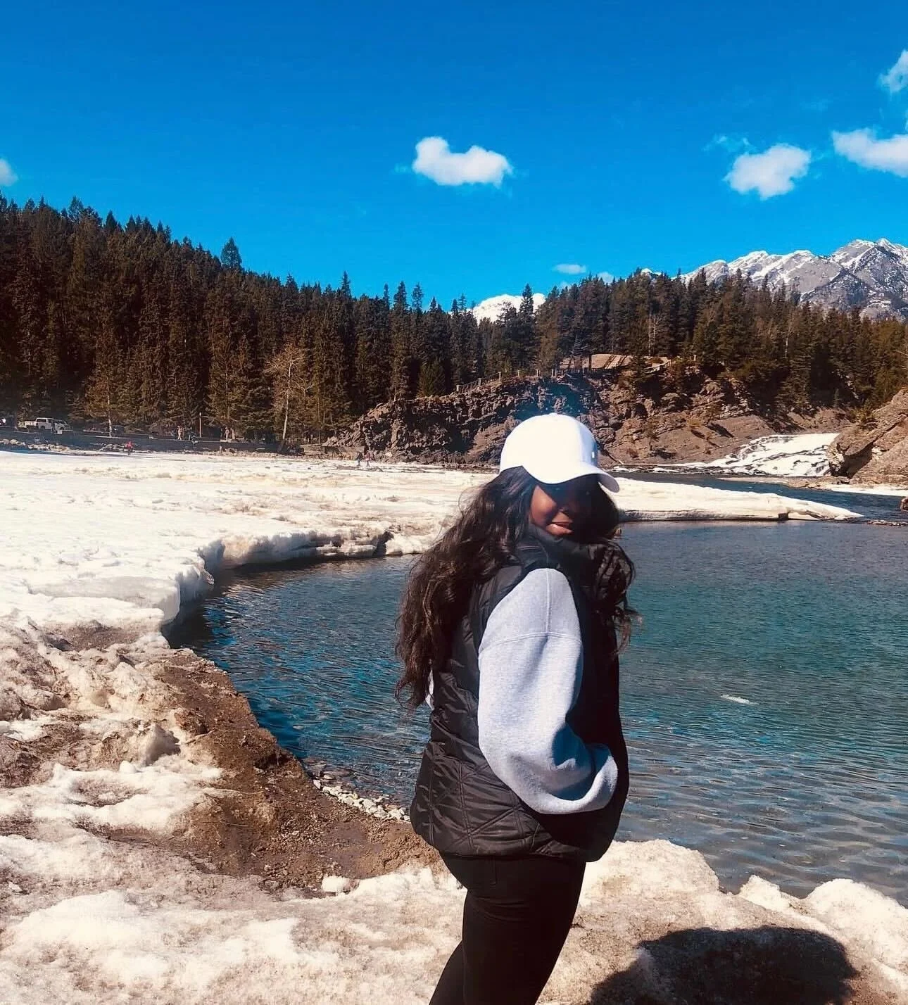 A woman standing near a body of water with snow, trees, and mountains in the background. She is wearing a white cap, sunglasses, a black vest over a gray hoodie, and black pants.