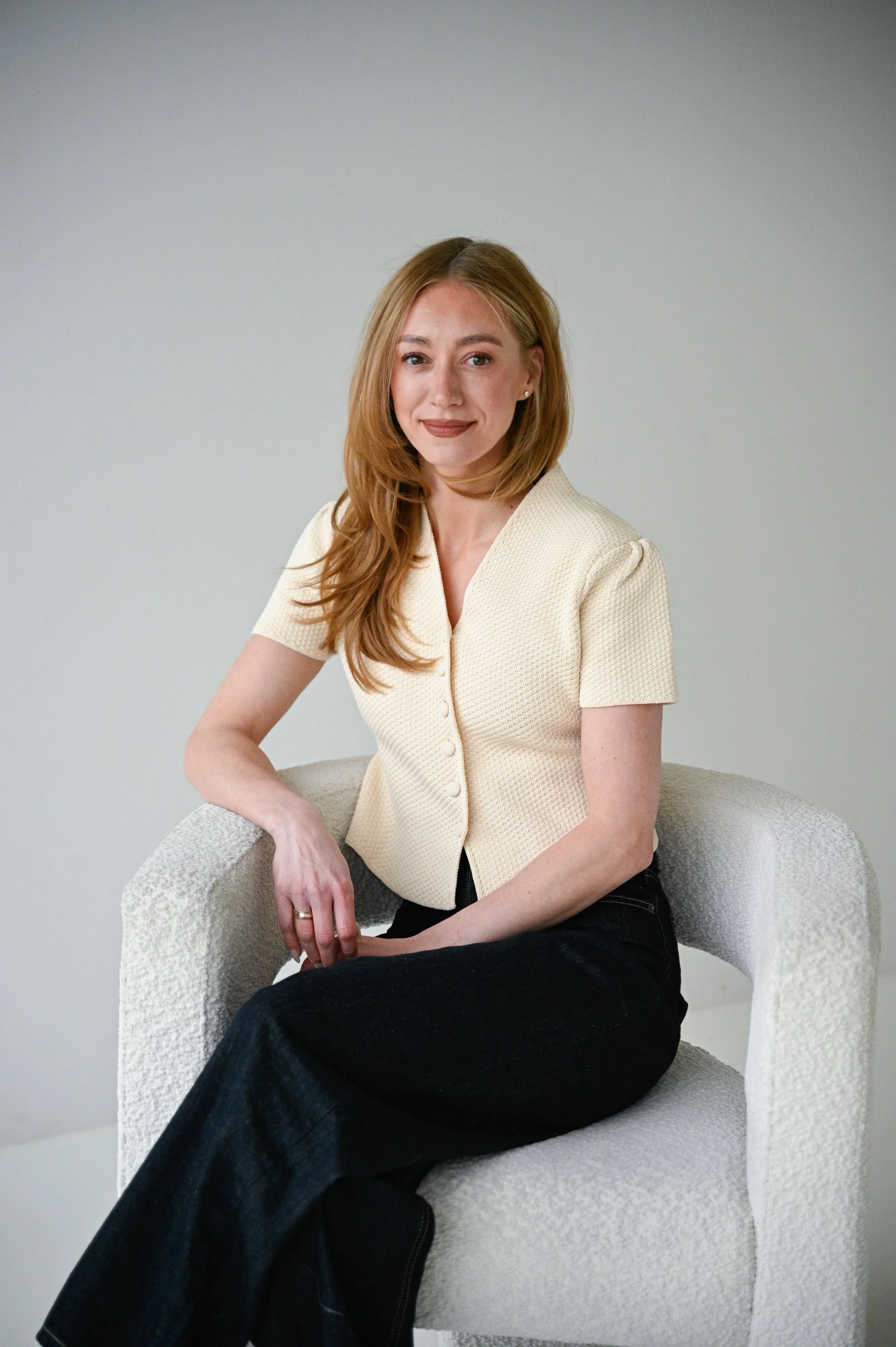 A woman with long red hair wearing a cream-colored short-sleeve top and dark pants, sitting in a white textured chair against a plain light background.