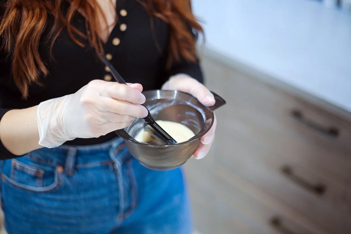 A person wearing white gloves and a black shirt is mixing a creamy substance in a black bowl with a black spoon.