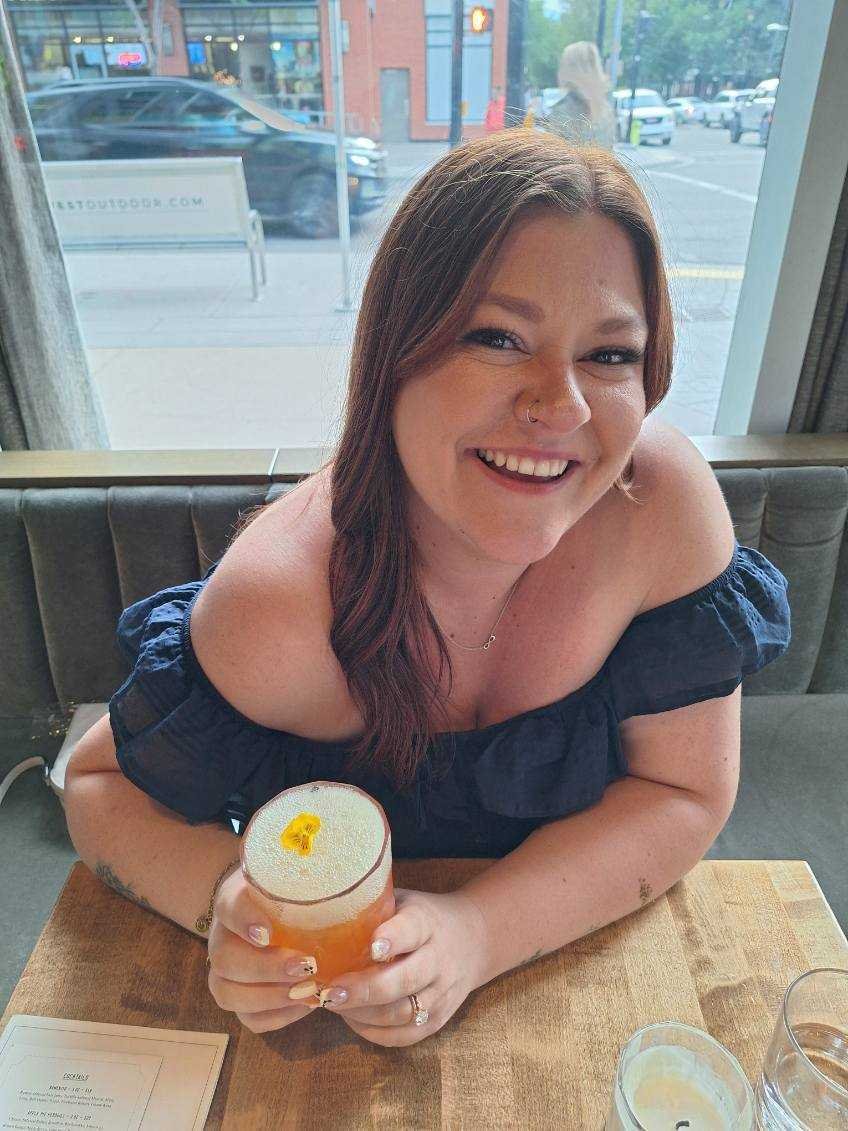 A woman with long brown hair sitting at a wooden table in a restaurant, smiling while holding a yellow-orange cocktail with a yellow flower garnish, near a large window with a city street view in the background.