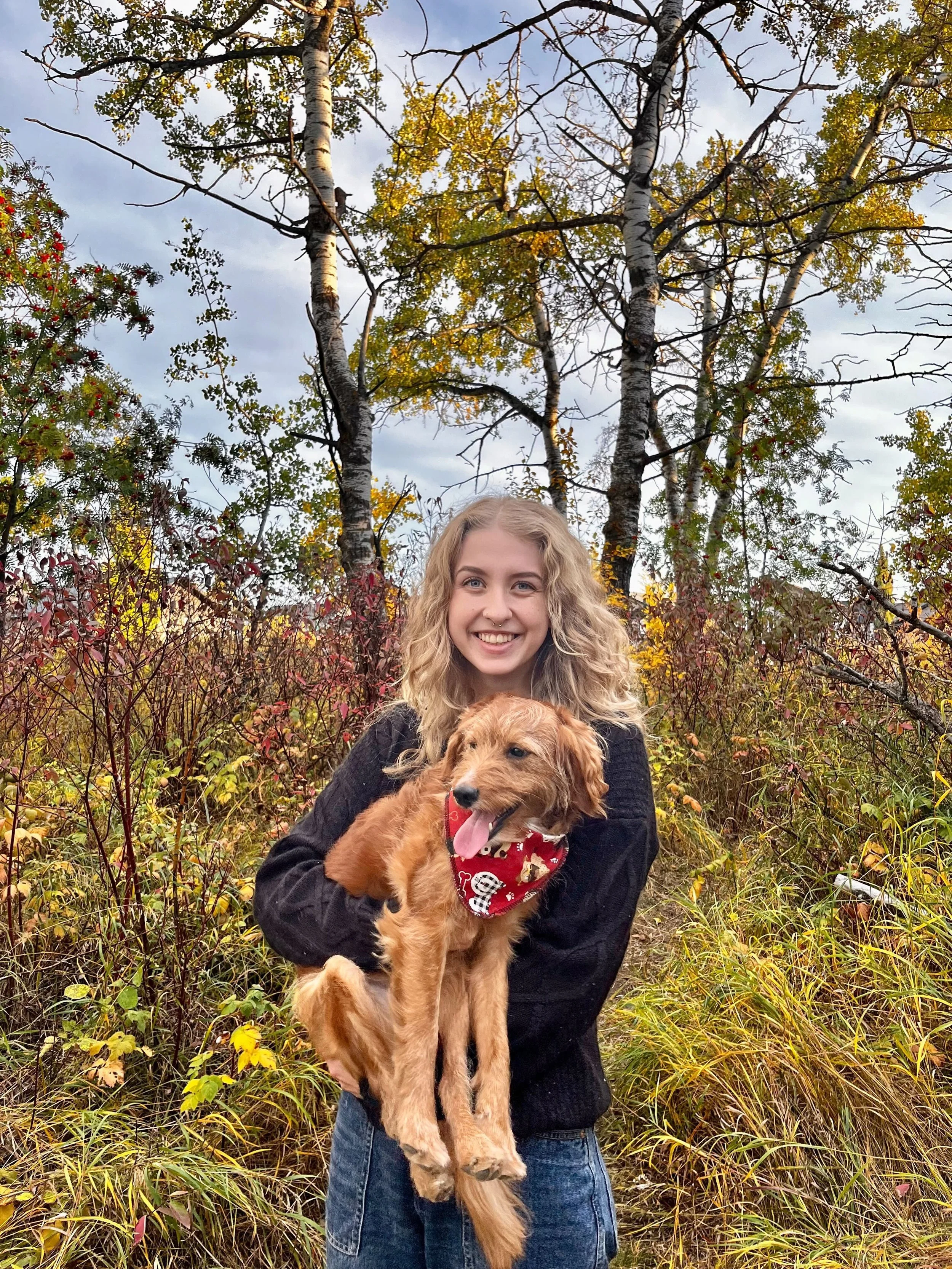 A young woman with curly blonde hair, wearing a black sweater and blue jeans, is smiling and holding a small, golden-brown dog with a red bandana, in an outdoor setting with autumn foliage and trees.