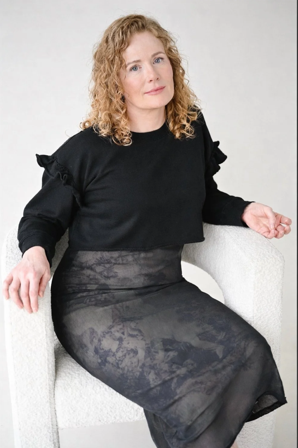 A woman with curly red hair sitting on a beige textured armchair, wearing a black top with ruffled shoulders and a black patterned skirt, against a plain light-colored background.