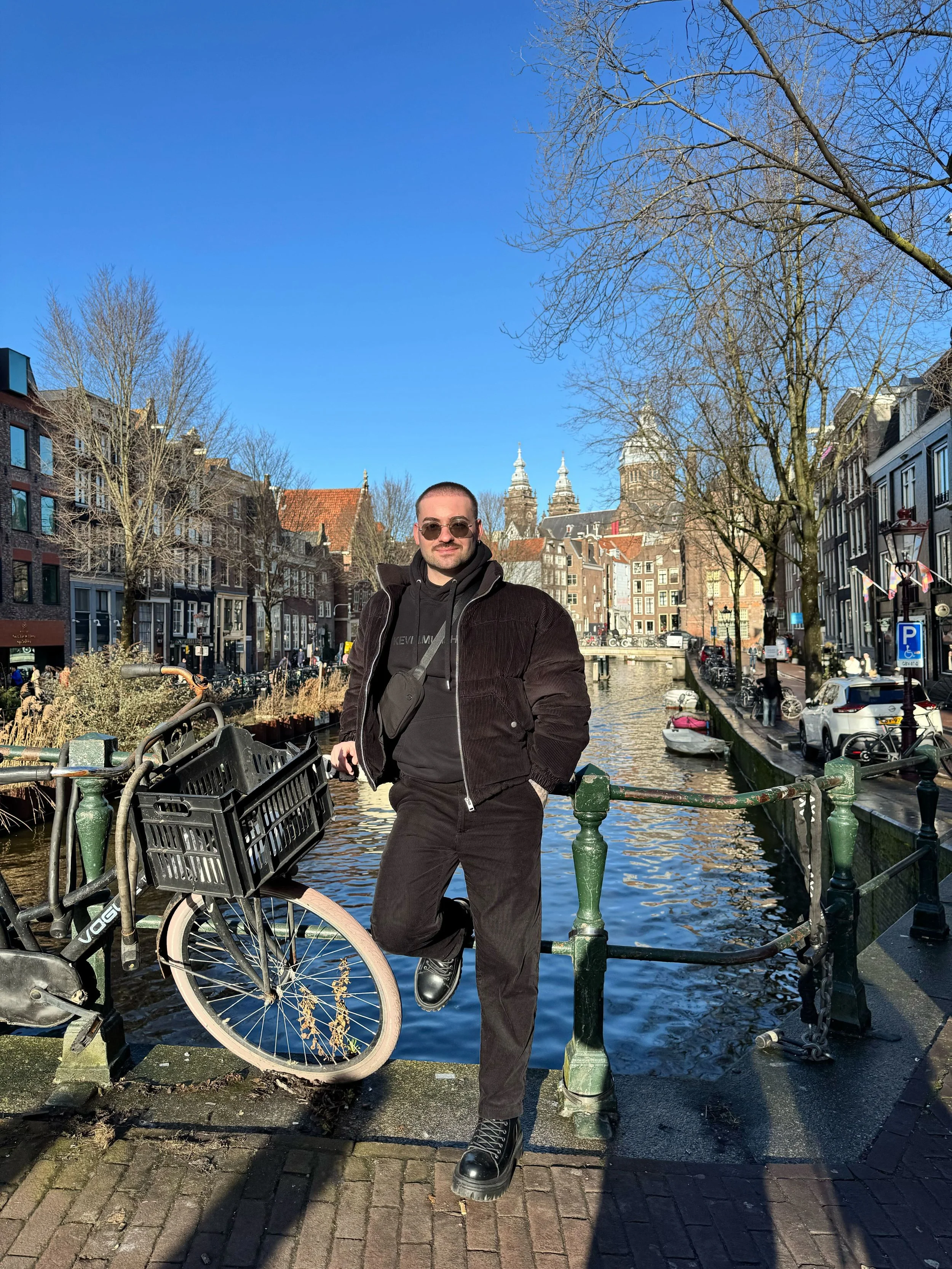 A man in black clothing and sunglasses stands on a bridge over a canal in a European city, with historic buildings and trees in the background.