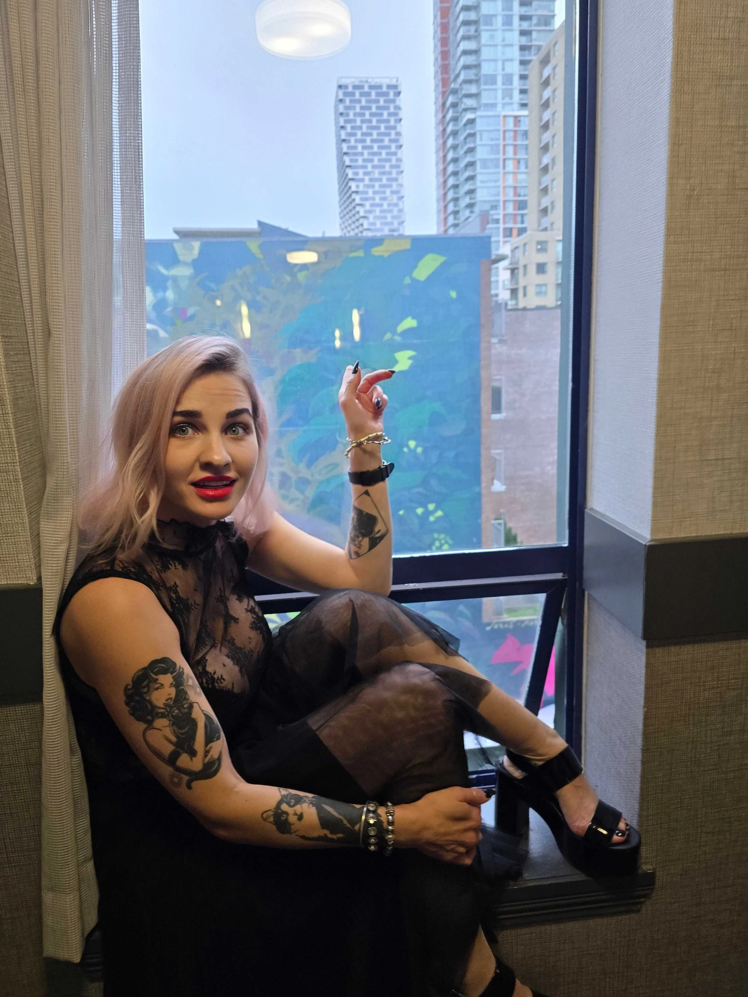 Young woman with blonde hair and tattoos sitting on a window seat, pointing at city buildings outside.