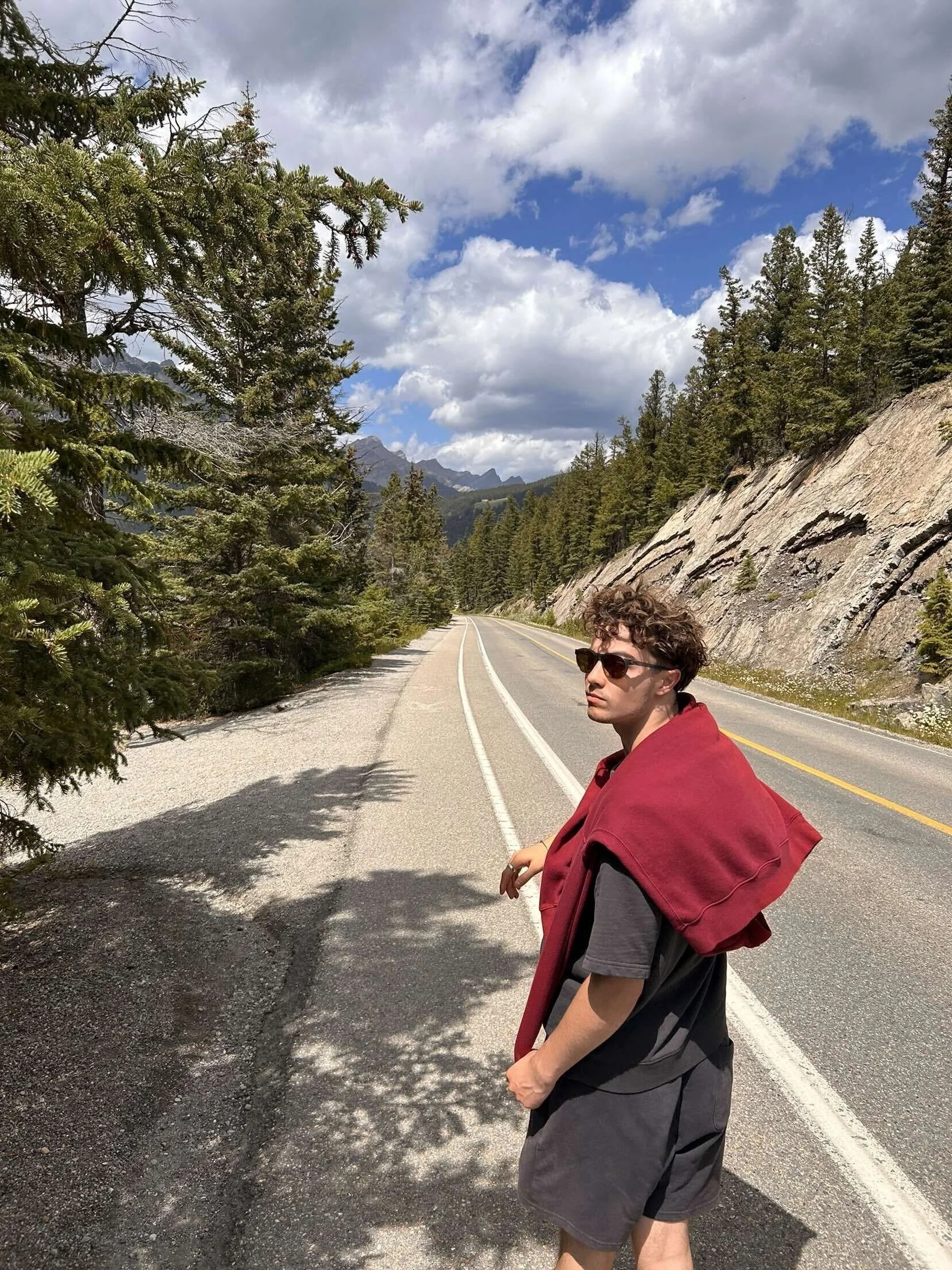 A young man with curly hair wearing sunglasses, a black t-shirt, and shorts, standing on the side of a mountain road, with a red jacket draped over his shoulders. He is looking back towards the camera with a scenic backdrop of tall pine trees, rocky cliffs, and a partly cloudy sky.