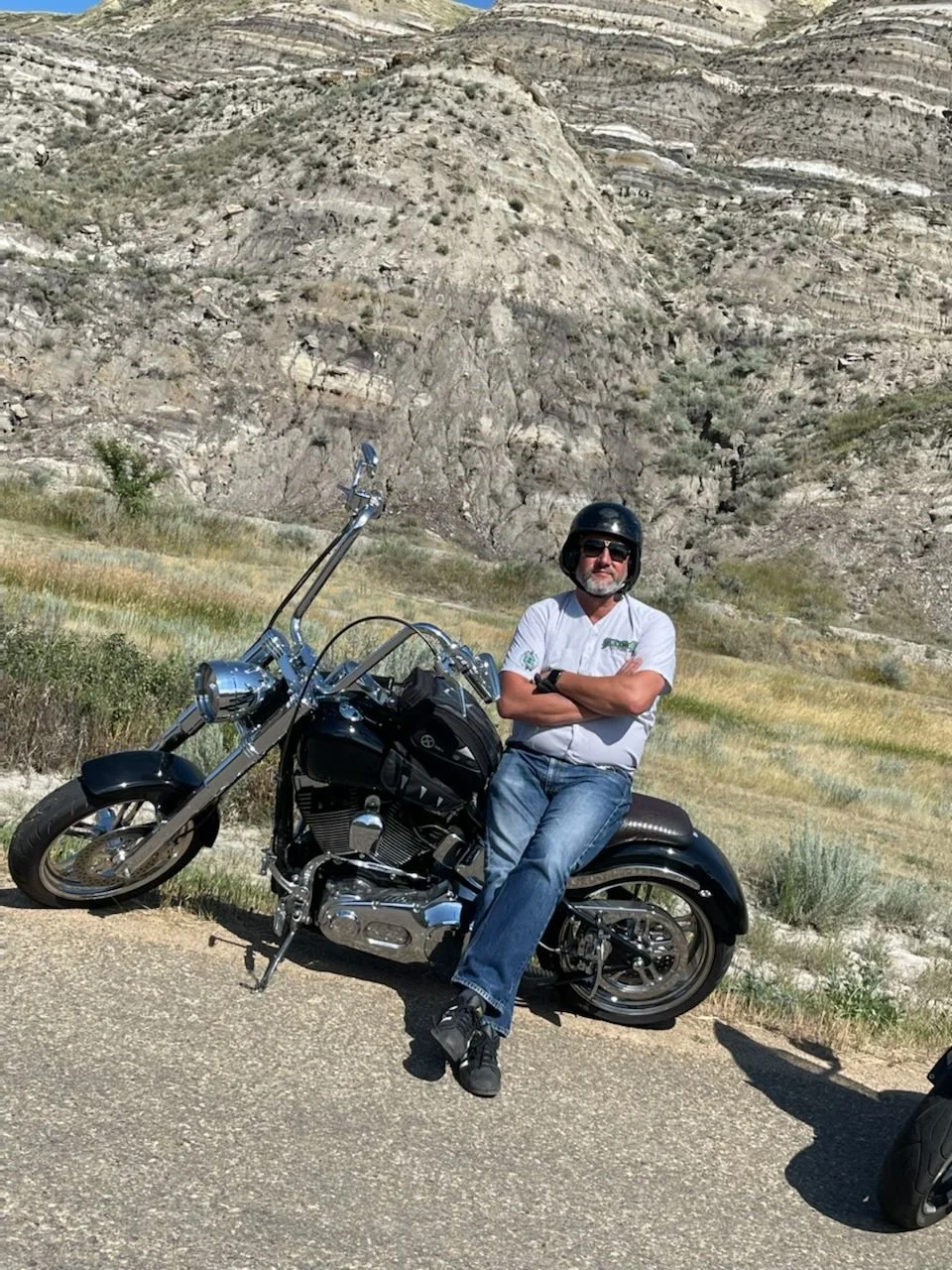 A man wearing a black helmet and sunglasses sits on a black motorcycle with his arms crossed, in front of a mountainous landscape with rocky terrain and sparse vegetation.
