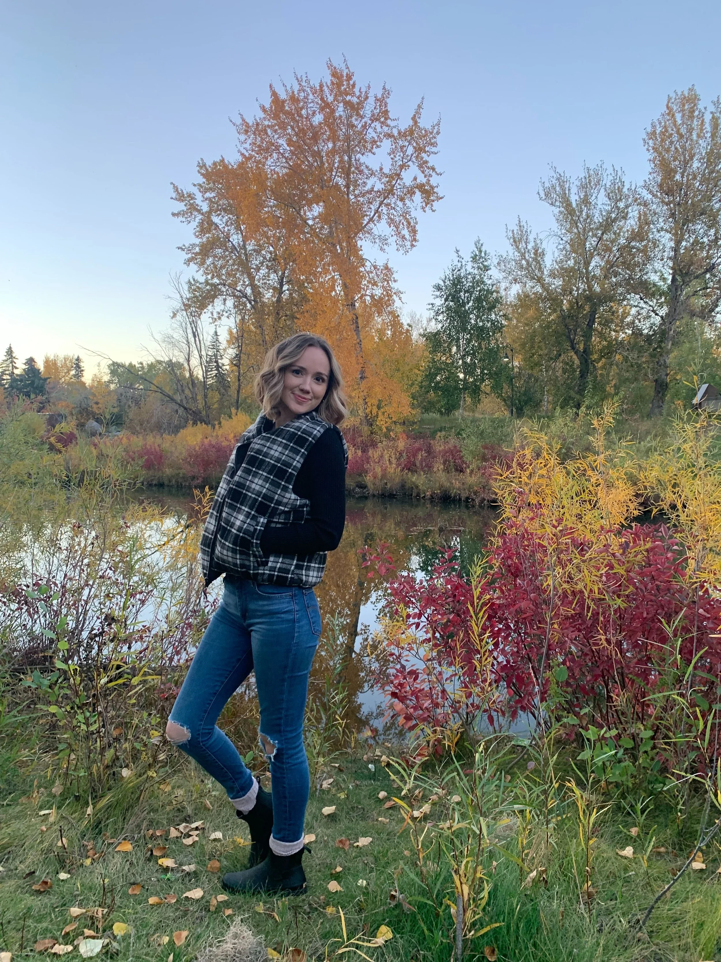 A woman standing outdoors near a body of water in autumn, surrounded by colorful trees and bushes.