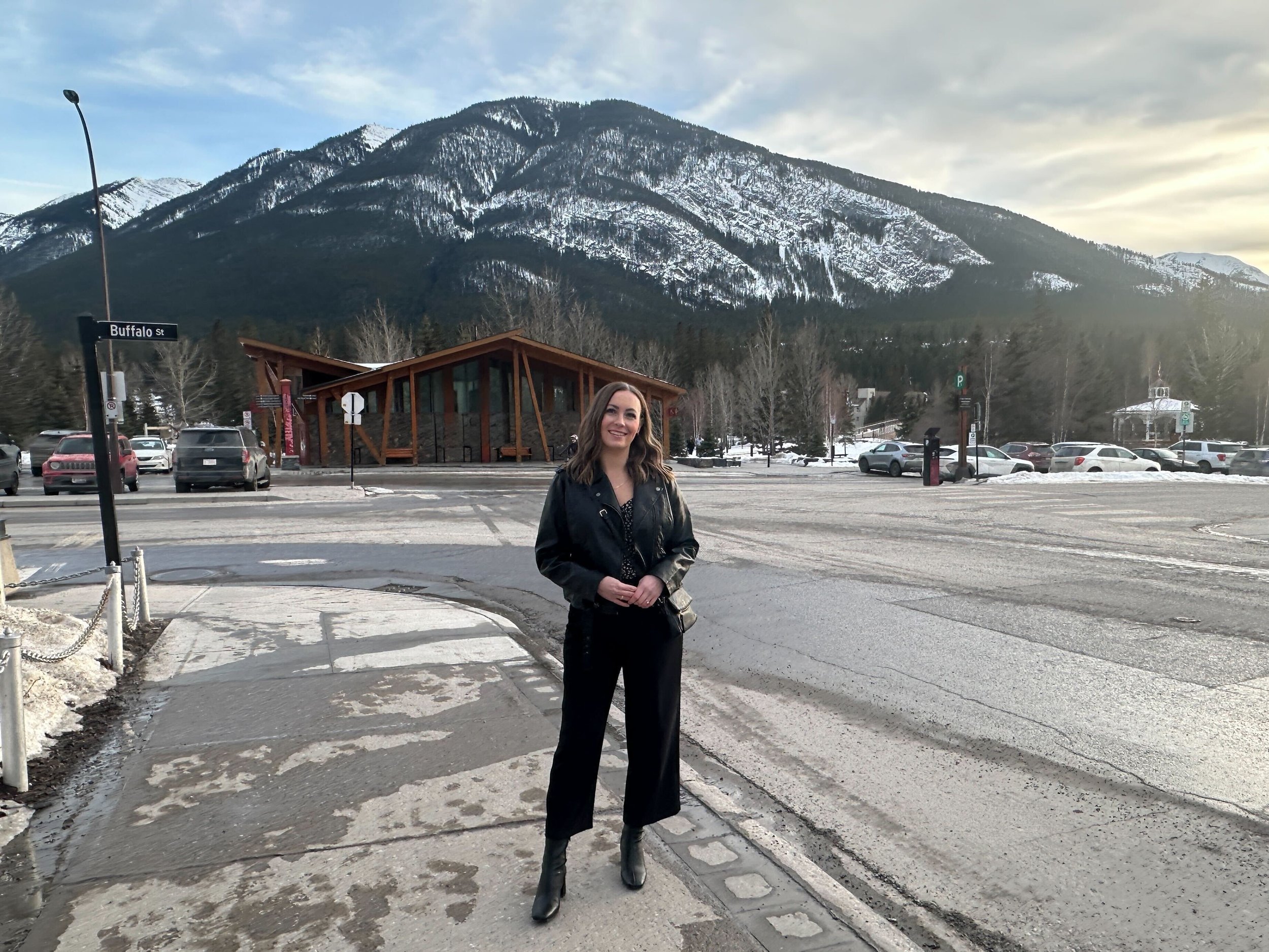A woman standing on a snow-dusted sidewalk in front of a parking lot and a modern wooden building with mountains in the background.