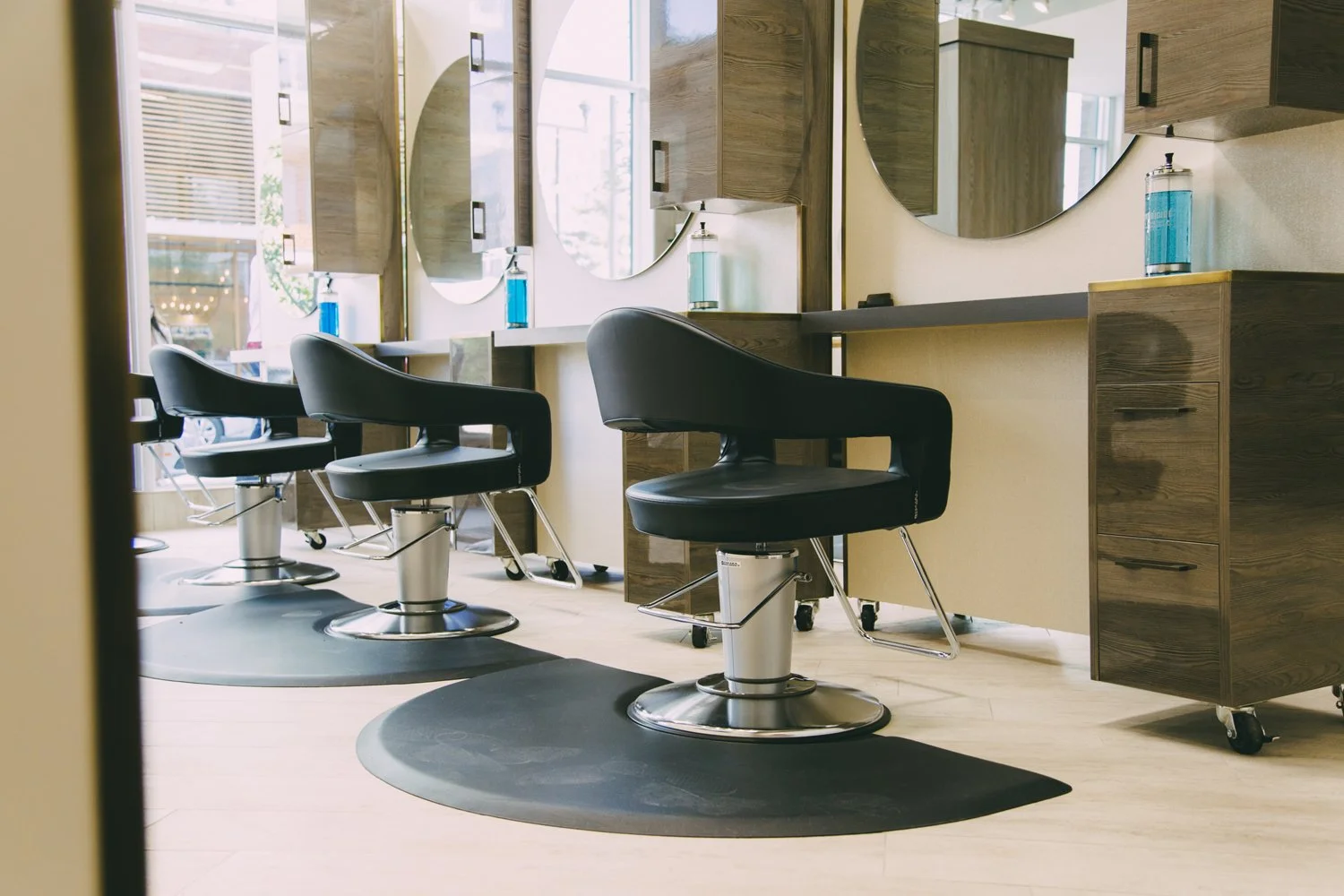 Empty hair salon with black styling chairs, large round mirrors, and wooden cabinets, illuminated by natural light through windows.