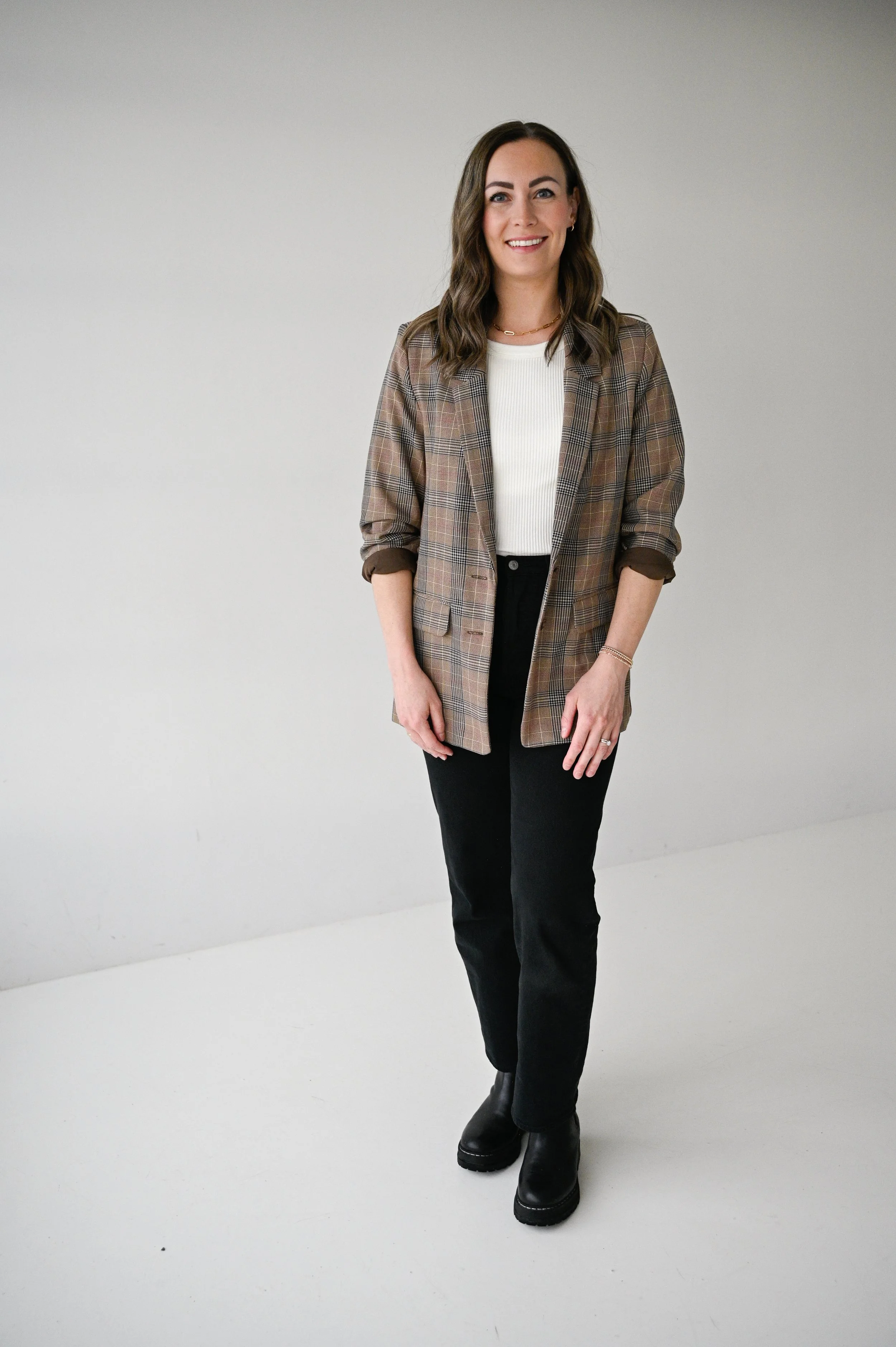 A woman with brown hair, wearing a brown plaid blazer, white top, black pants, and black boots, standing and smiling against a plain light gray background.