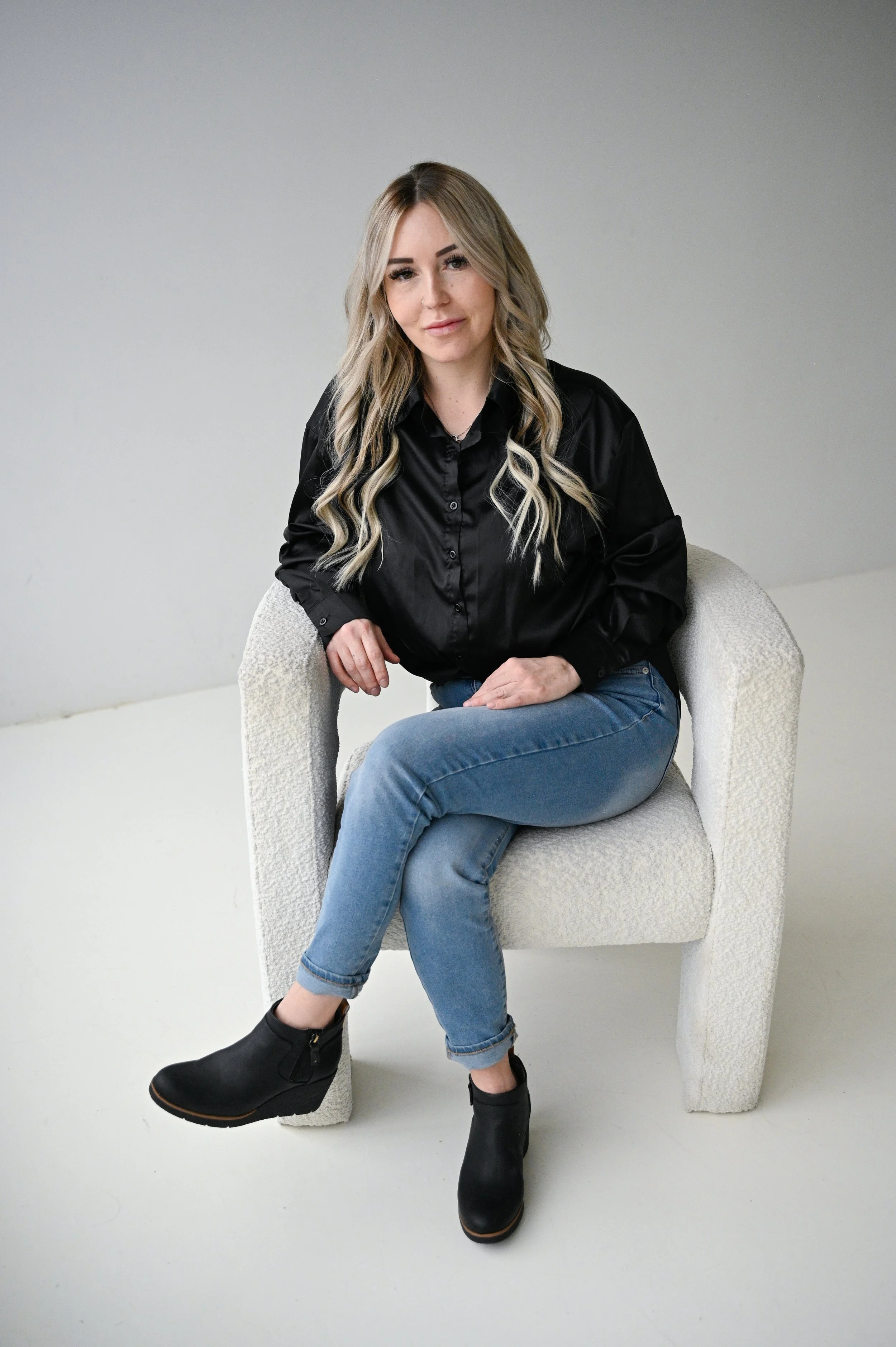 A woman with long blonde hair, wearing a black shirt, blue jeans, and black ankle boots, sitting on a textured white armchair against a plain white background.