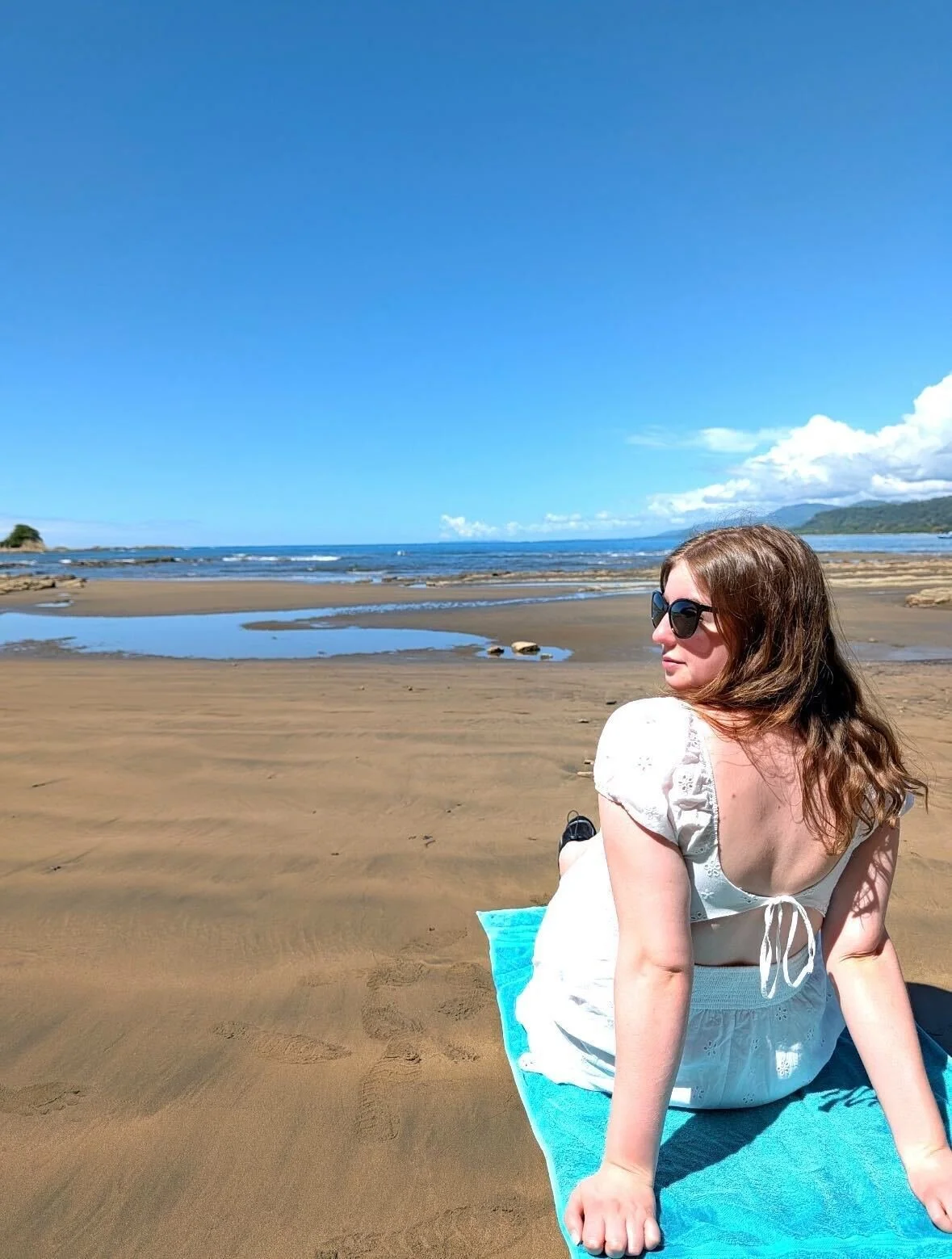 A woman with brown hair, wearing sunglasses and a white dress, sitting on a blue towel on a sandy beach with water and mountains in the background under a partly cloudy blue sky.