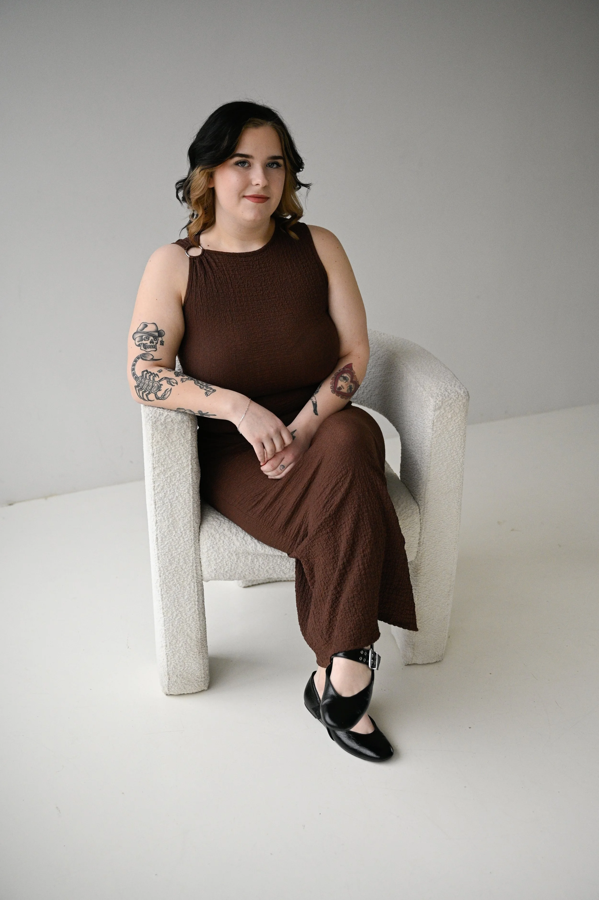 A woman with black and blonde hair sitting on a white textured armchair in a minimalistic room with a plain gray wall. She is wearing a brown sleeveless dress, black shoes, and has tattoos on her arms.