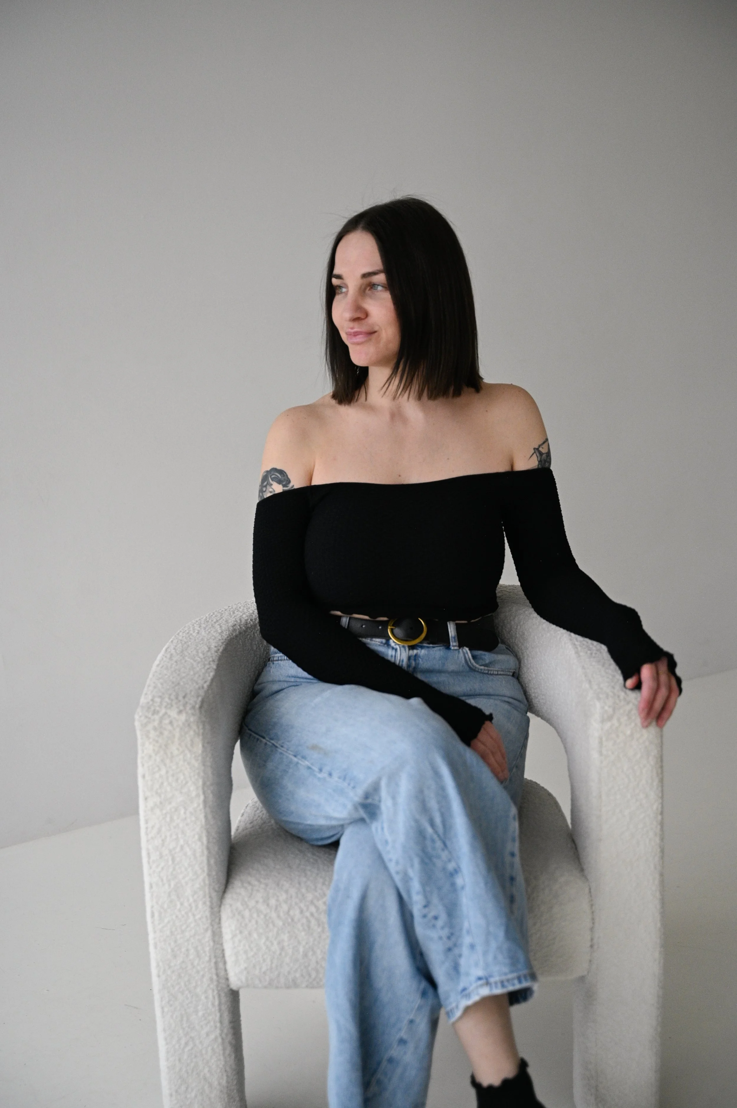 A young woman with black hair and tattoos, wearing an off-the-shoulder black top and light blue jeans, sitting on a white textured armchair against a plain light gray background.