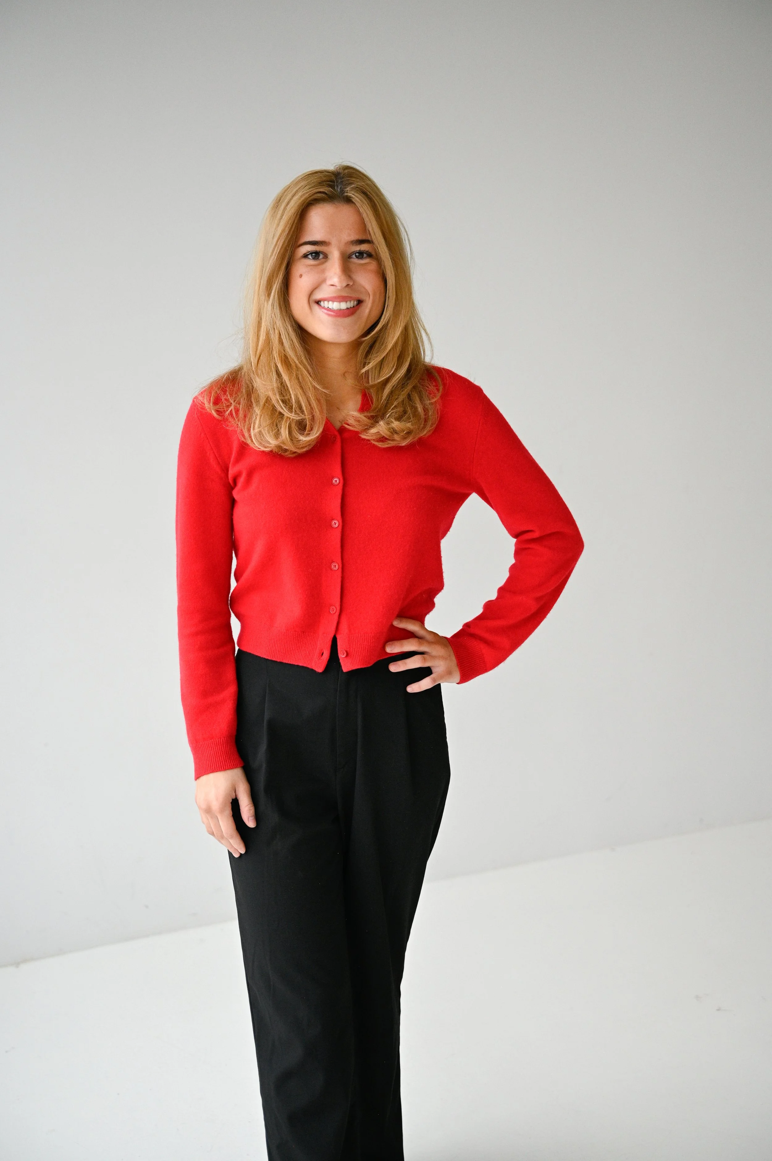 A young woman with blonde hair wearing a red cardigan and black pants, standing against a plain white background, smiling with one hand on her hip.