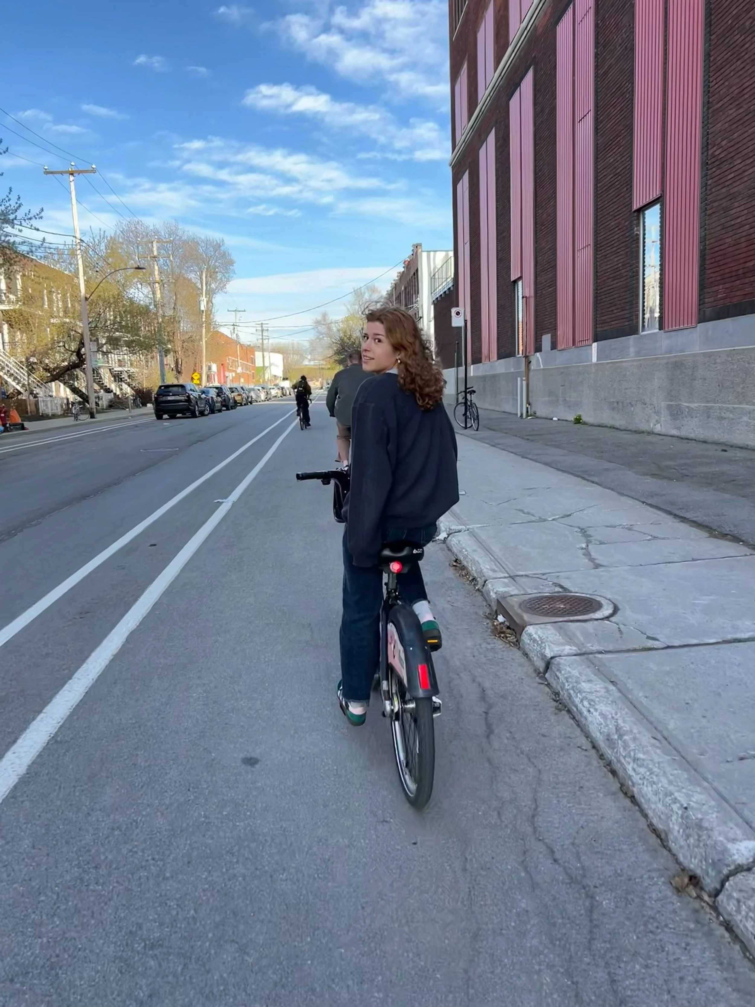A young woman riding a bicycle on a city street with cars parked along the side. She has curly hair and is wearing a dark jacket. The sky is partly cloudy with some blue visible, and the street is lined with buildings, including a large building with pink panels.