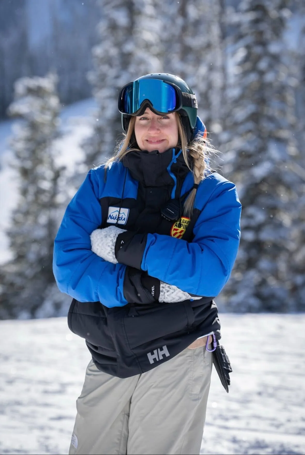 A woman in snow gear standing outdoors in a snowy landscape, wearing a blue jacket, black gloves, white mittens, and a helmet with goggles, smiling with her arms crossed.