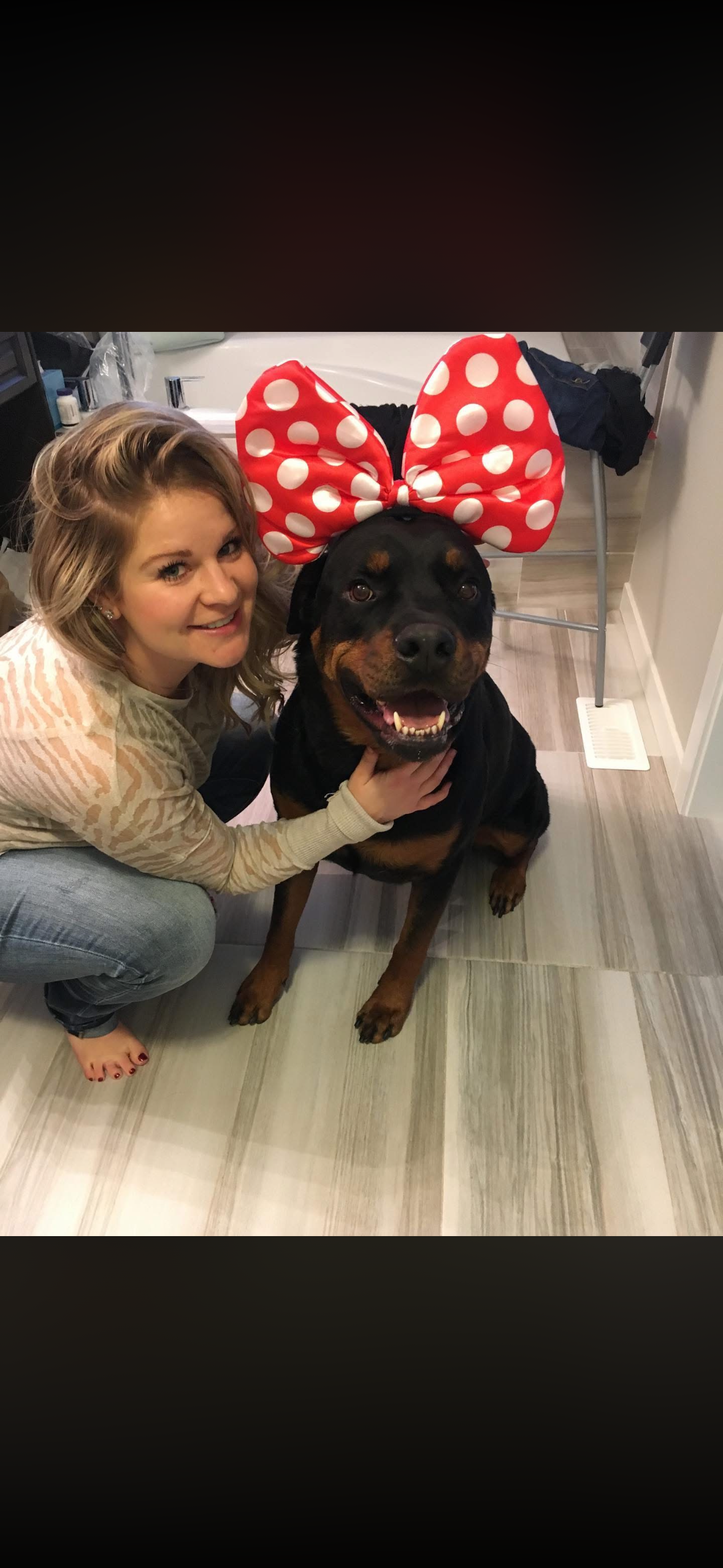 A woman with blonde hair crouches next to a large Rottweiler wearing a red and white polka dot bow. Both are smiling in a room with patterned tiled flooring, a small vent, and various household items in the background.