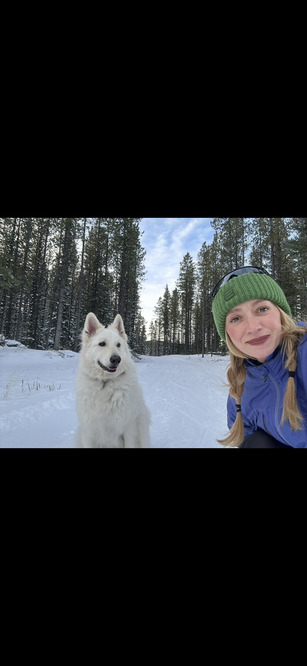 A woman and a white dog outdoors in a snowy forest, smiling at the camera. The woman is wearing a green beanie, sunglasses, and a blue jacket. The dog appears to be a Samoyed, standing on the snow-covered ground.”}