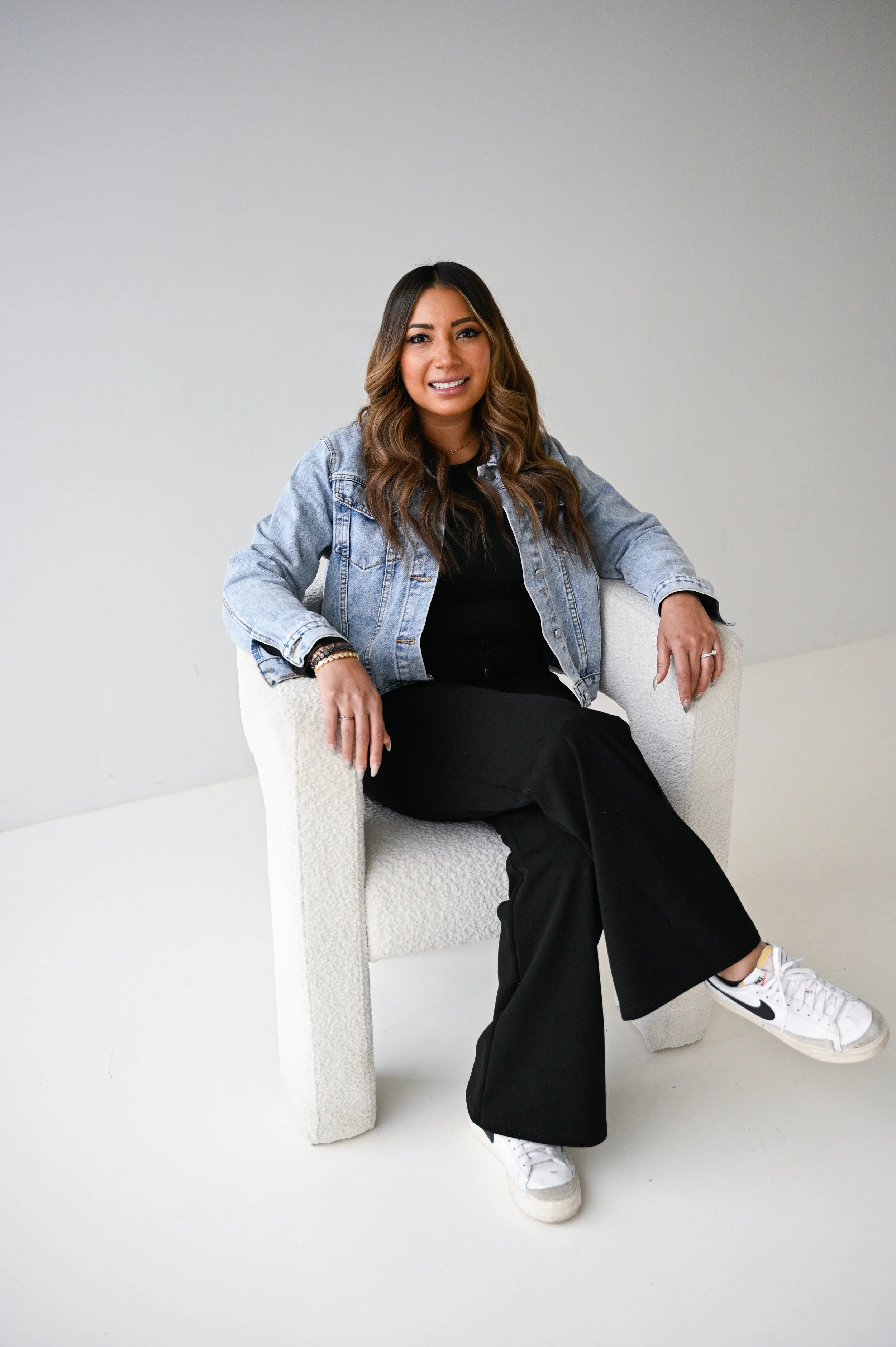 A woman sitting on a textured white chair against a plain light grey background, wearing a denim jacket, black pants, white sneakers, and smiling at the camera.
