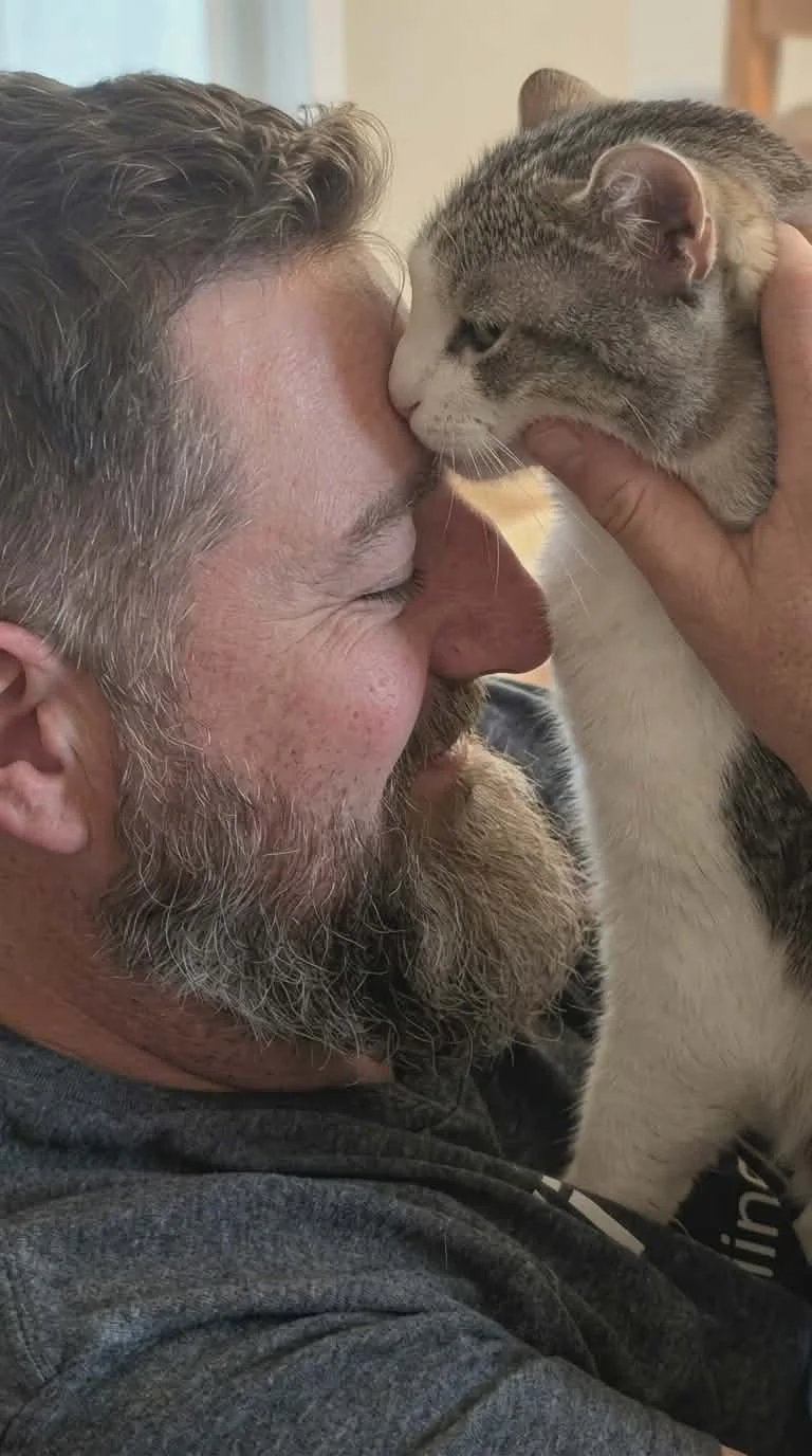 A man with gray hair and a beard is holding a gray and white tabby cat close to his face, touching noses in a tender moment.