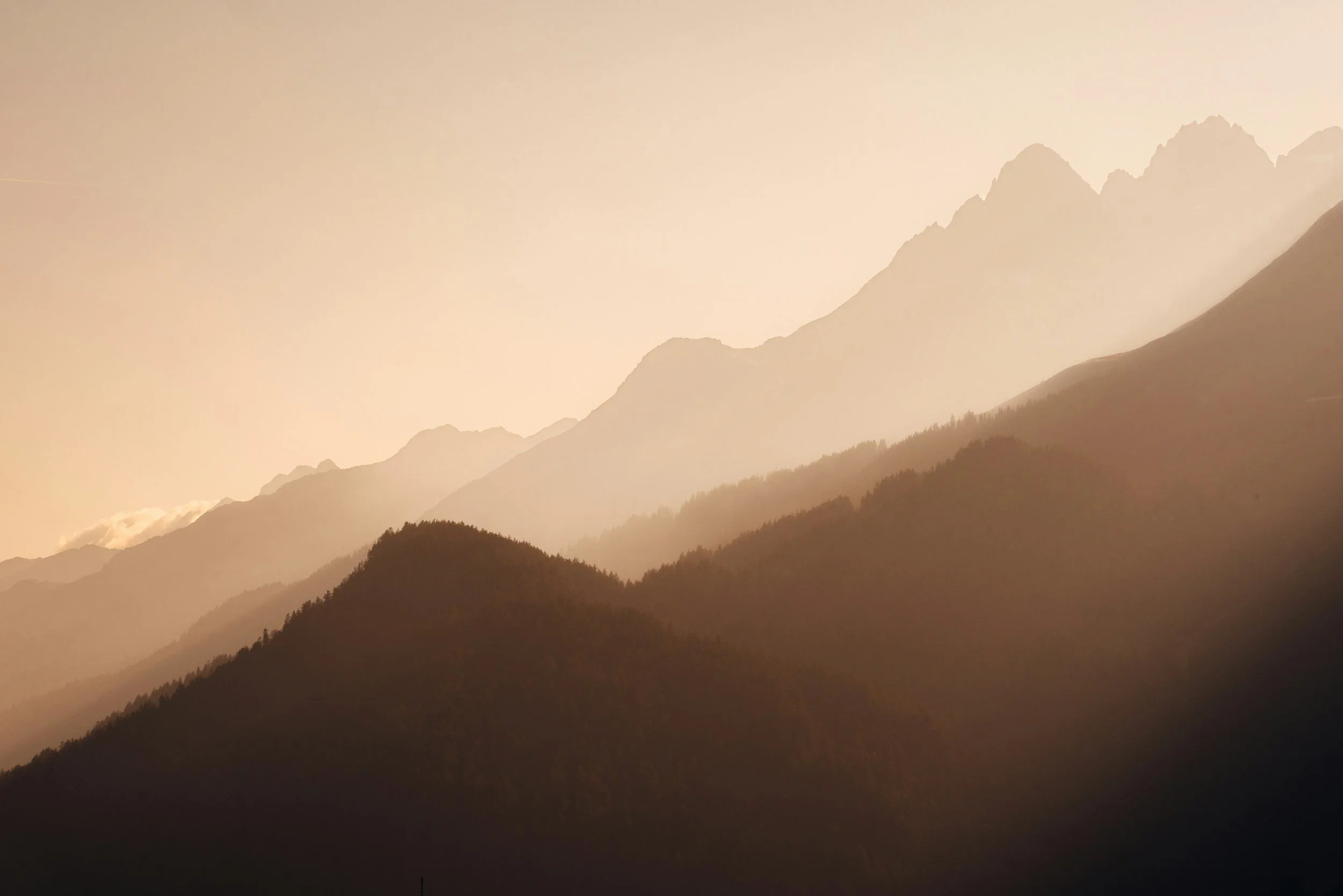 Sunset over mountain range with silhouettes of forested hills in the foreground.